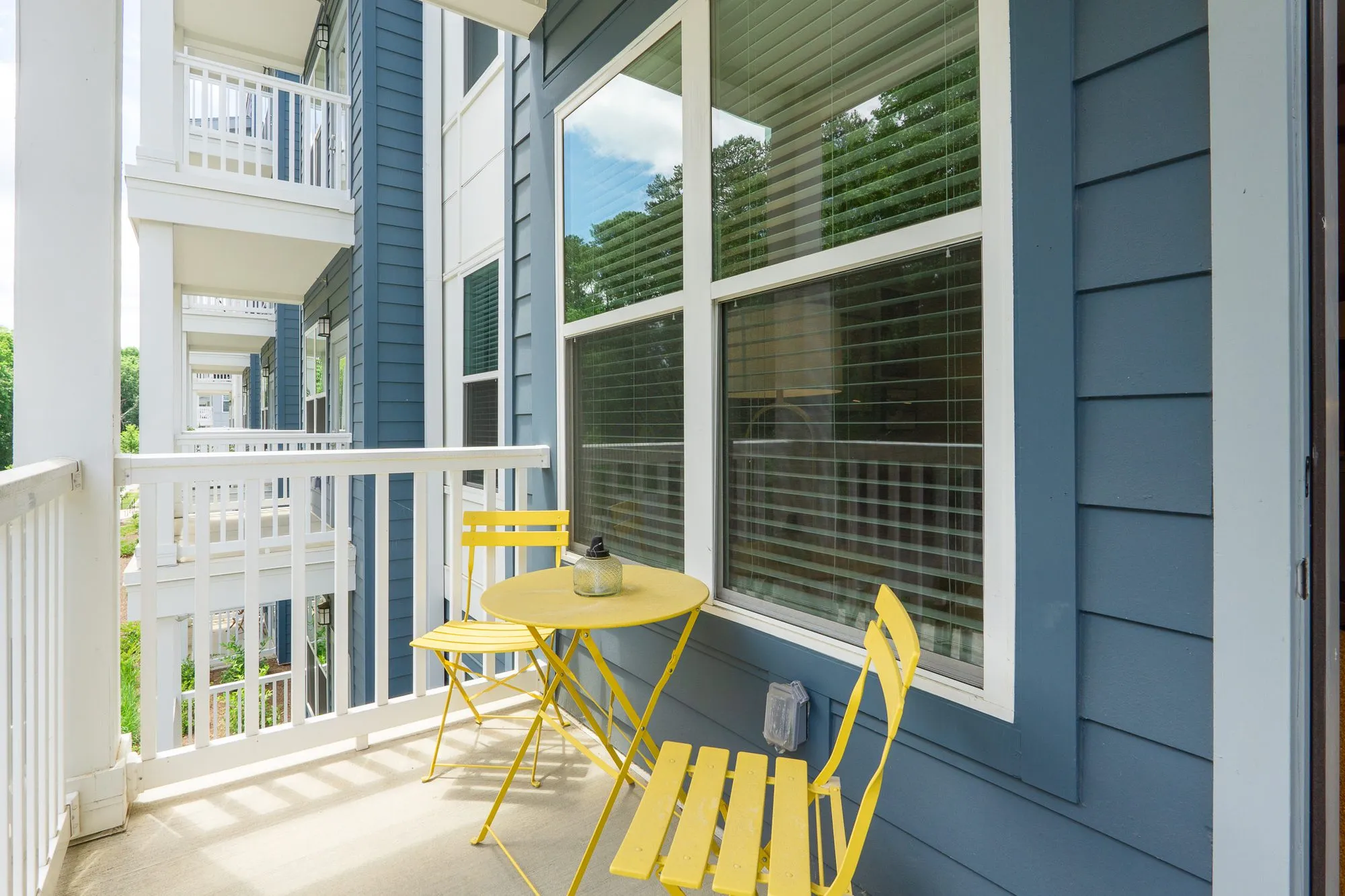 Multifamily apartment balcony with a yellow folding bistro table and chairs beside blue siding and a white railing
