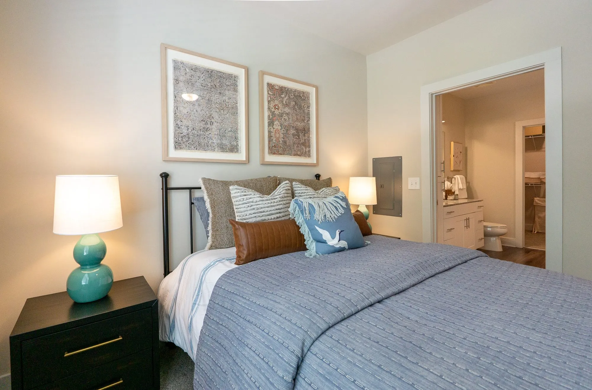 Multifamily apartment bedroom with an iron bed, blue coverlet and bird pillow between a teal lamp and two textile prints