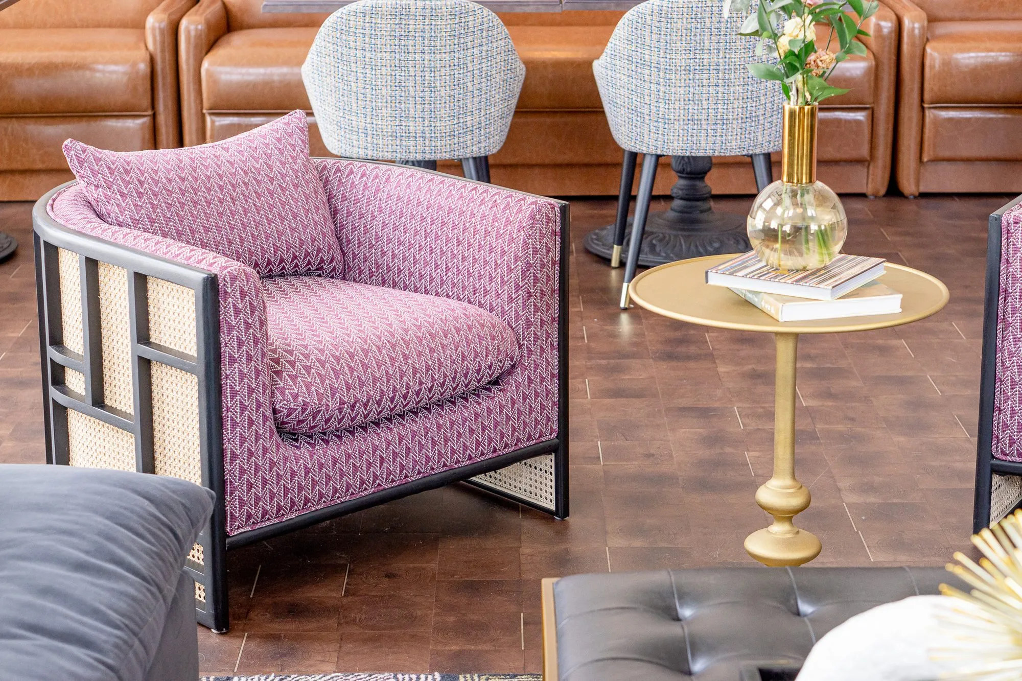 Multifamily amenity clubroom with a pink patterned cane armchair beside a brass side table, grey chairs and leather booths