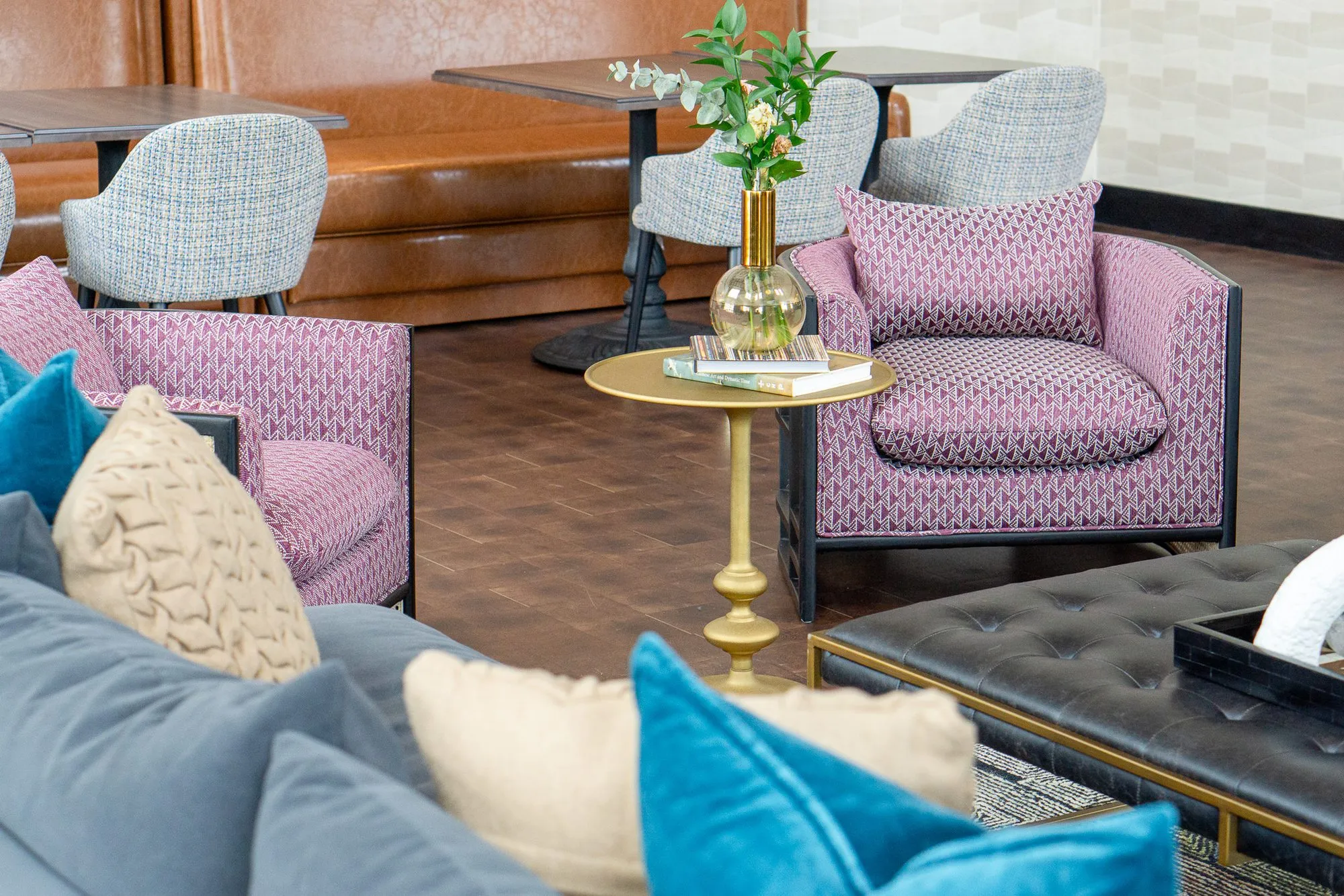 Multifamily amenity clubroom with pink armchairs around a brass side table, a navy sofa with teal pillows and booths beyond