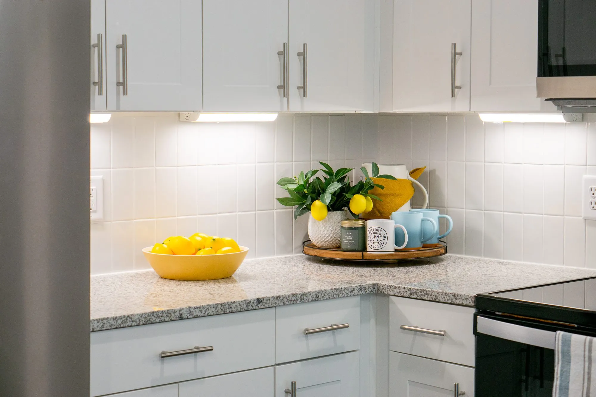 Multifamily apartment model kitchen corner with white cabinets, a granite counter, a bowl of lemons and mugs on a tray