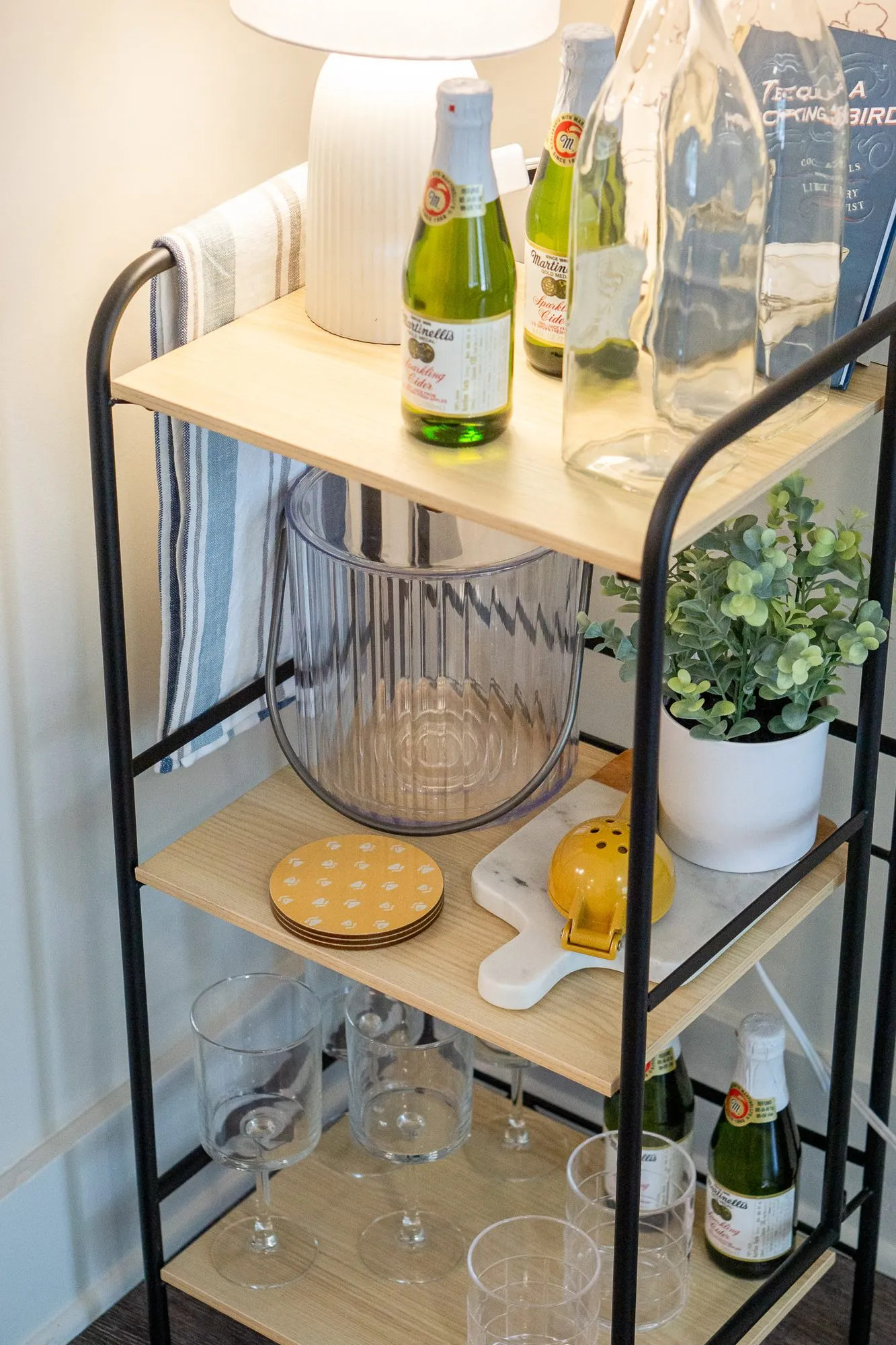 Multifamily interior detail: black metal bar cart with wood shelves holding sparkling wine bottles, glasses and a plant