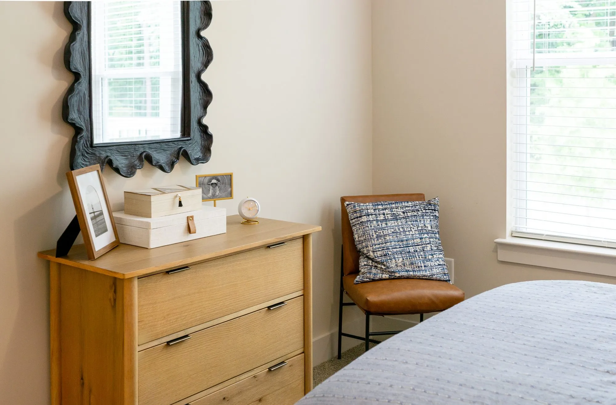 Multifamily apartment bedroom corner with a wood dresser under a black scalloped mirror, a leather chair by the window