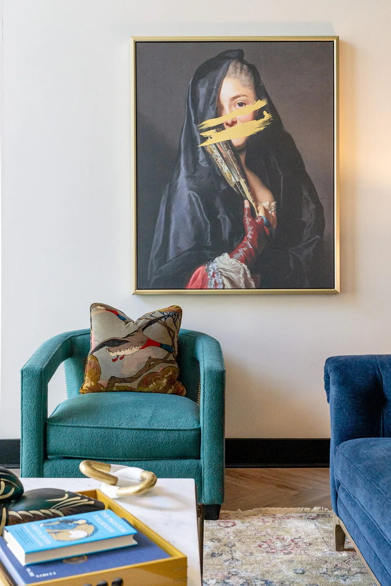 Multifamily amenity lobby lounge with a teal armchair and bird pillow beneath the veiled portrait, a navy sofa arm at right