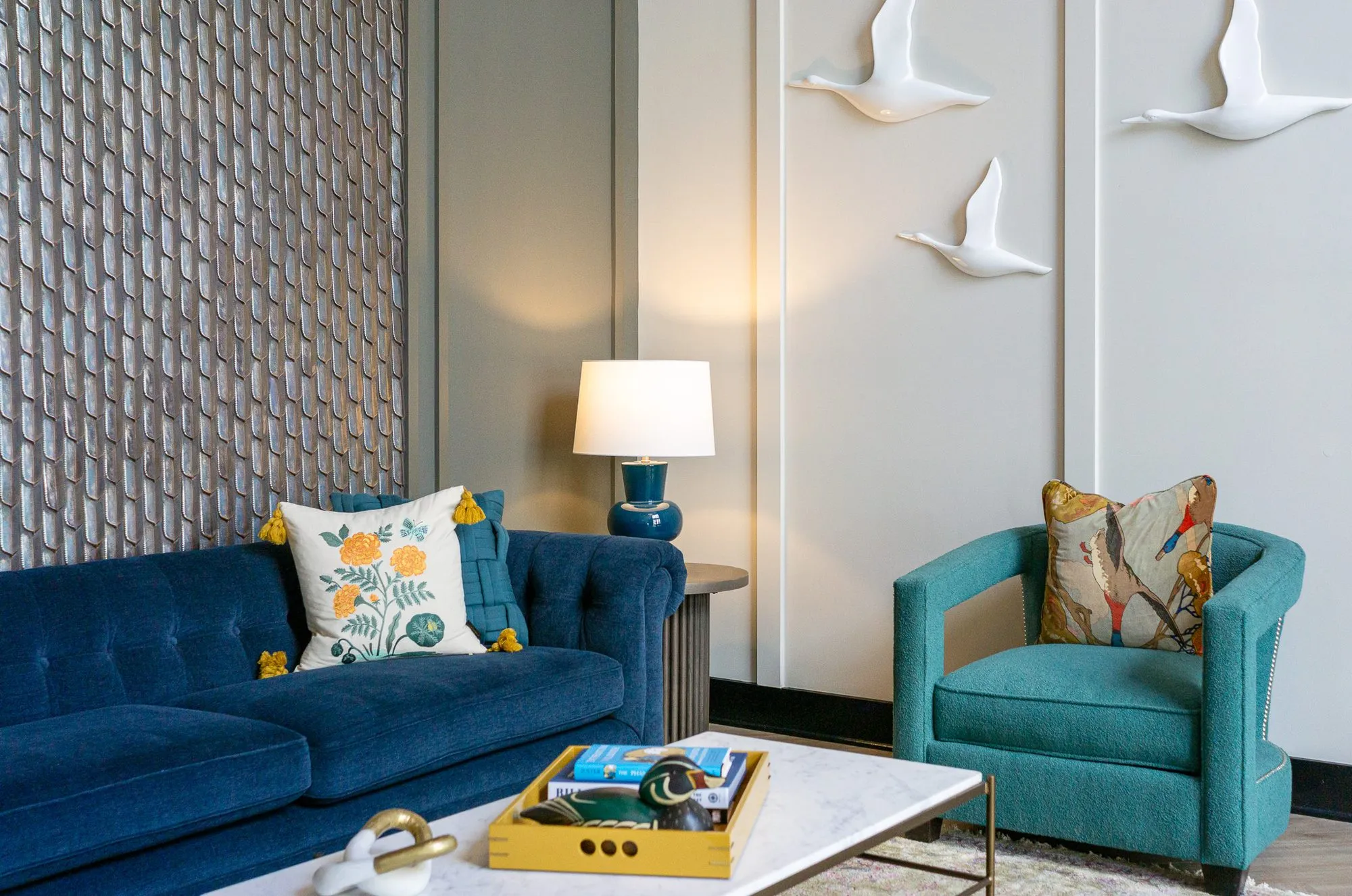 Multifamily amenity lobby lounge with a navy tufted sofa and teal armchair, white bird sculptures across the panelled wall