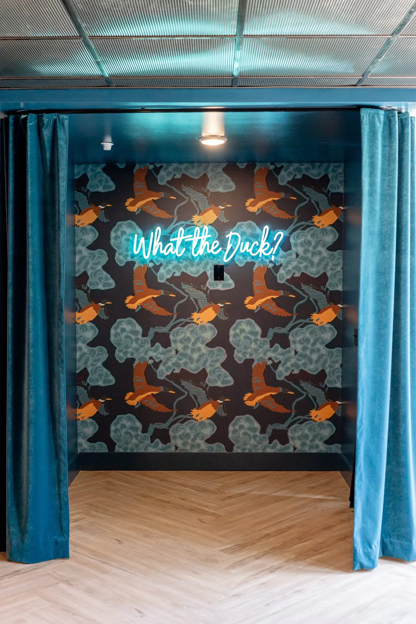 Multifamily amenity alcove seen through teal velvet curtains to a neon What the Duck? sign on duck-print wallpaper