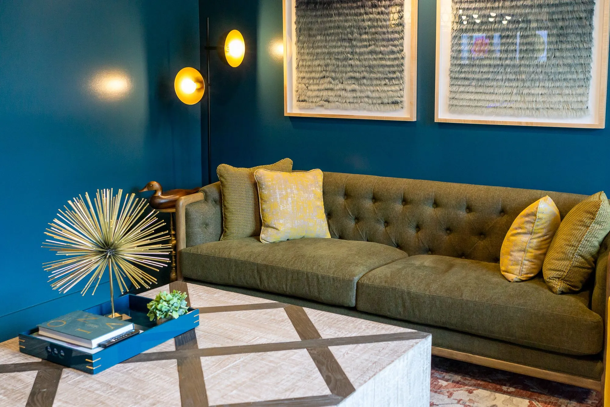 Multifamily interior detail: olive tufted sofa with yellow pillows on a dark teal wall under two textural artworks