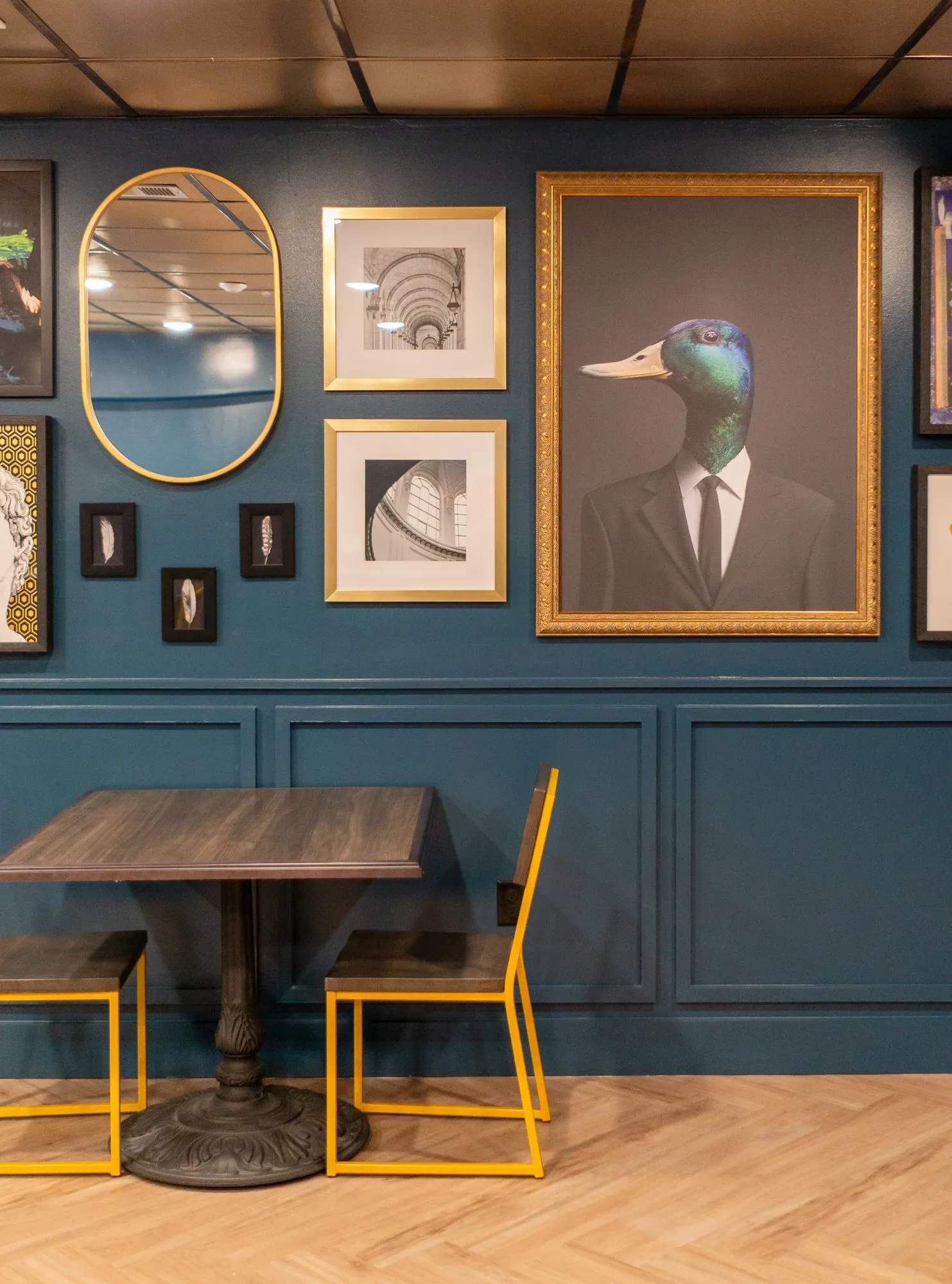 Multifamily amenity gallery wall on teal panelling with a duck-headed gentleman portrait and oval mirror above yellow chairs