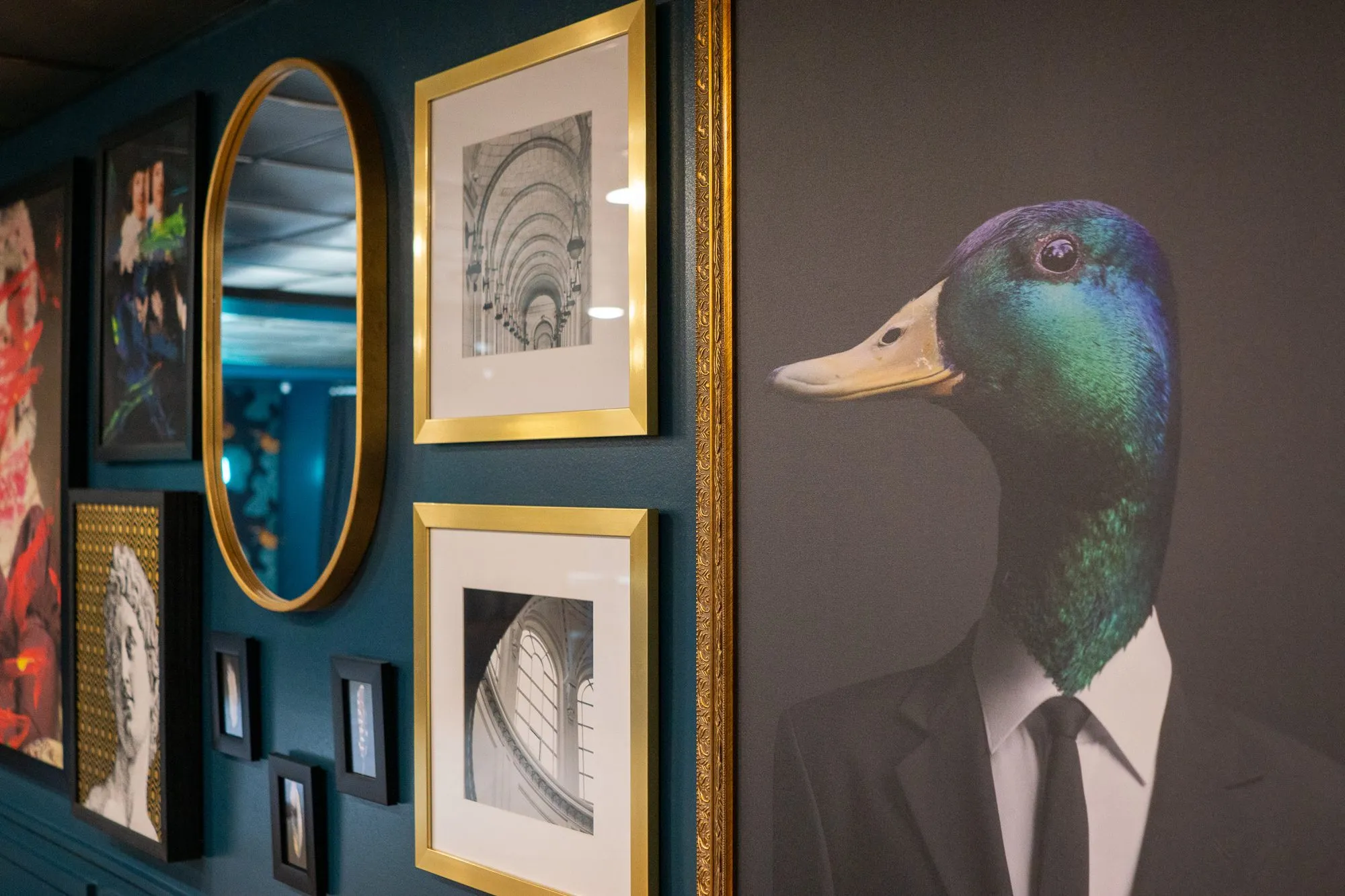 Multifamily amenity gallery wall with a duck-headed gentleman portrait in a gilt frame beside an oval mirror