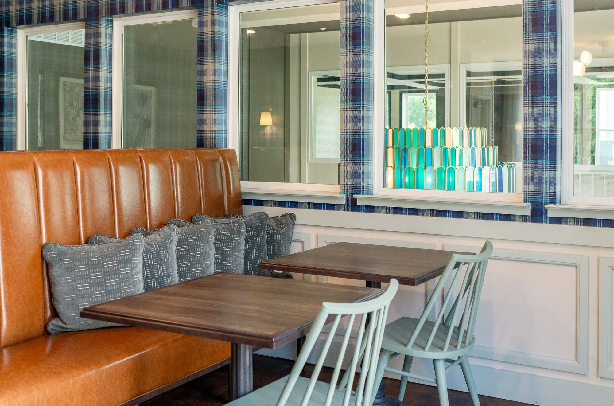Multifamily amenity cafe with a cognac banquette, grey pillows and wood tables with sage spindle chairs, plaid walls