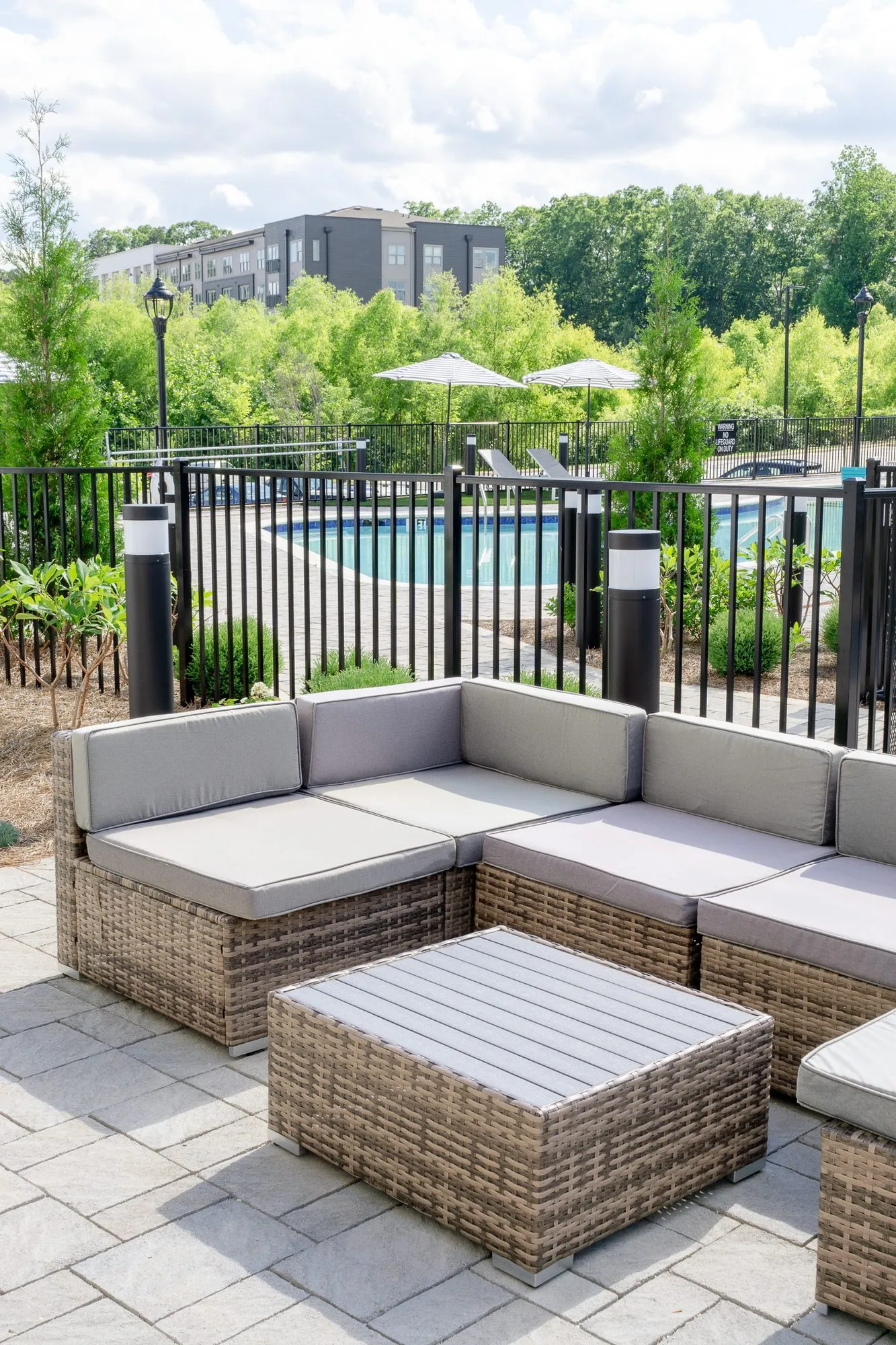 Multifamily community patio with a wicker sectional and slatted table, the pool and trees beyond the fence