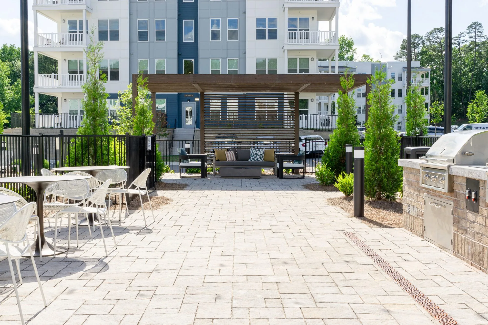 Multifamily community courtyard with white metal dining chairs, a built-in grill and a timber pergola before the apartments