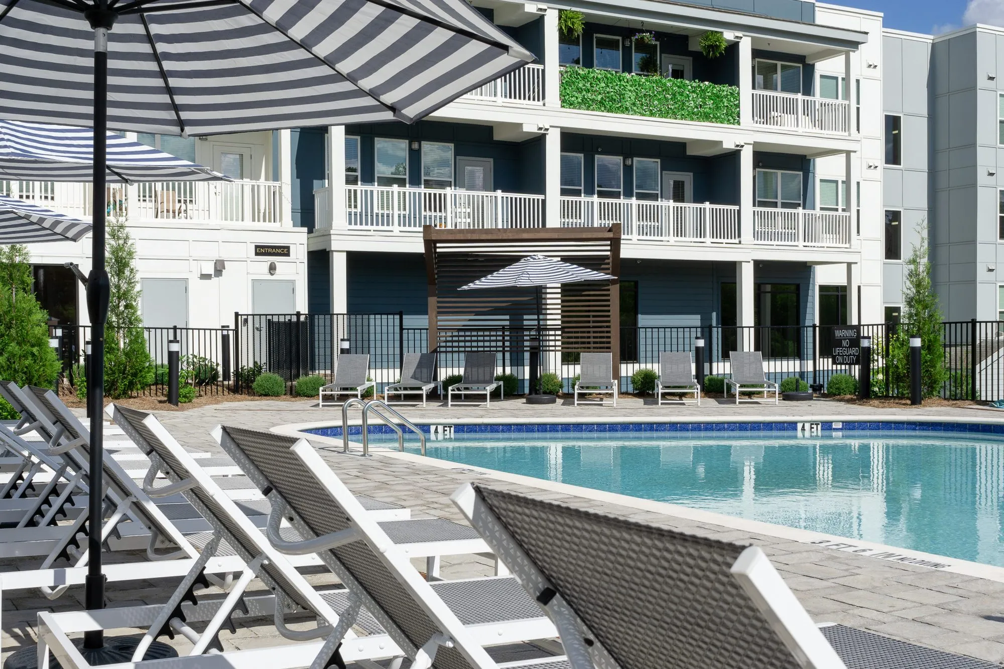 Multifamily community pool with grey and white chaises along the deck, a timber pergola and three storey apartments beyond