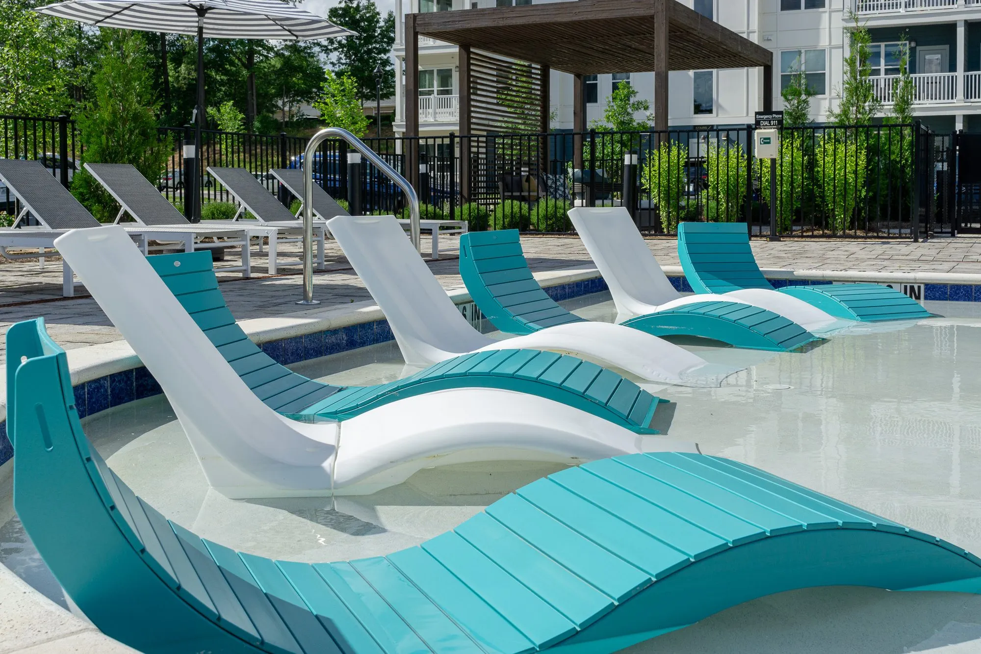 Multifamily community pool with teal and white curved loungers in the shallow shelf, a pergola and apartments beyond