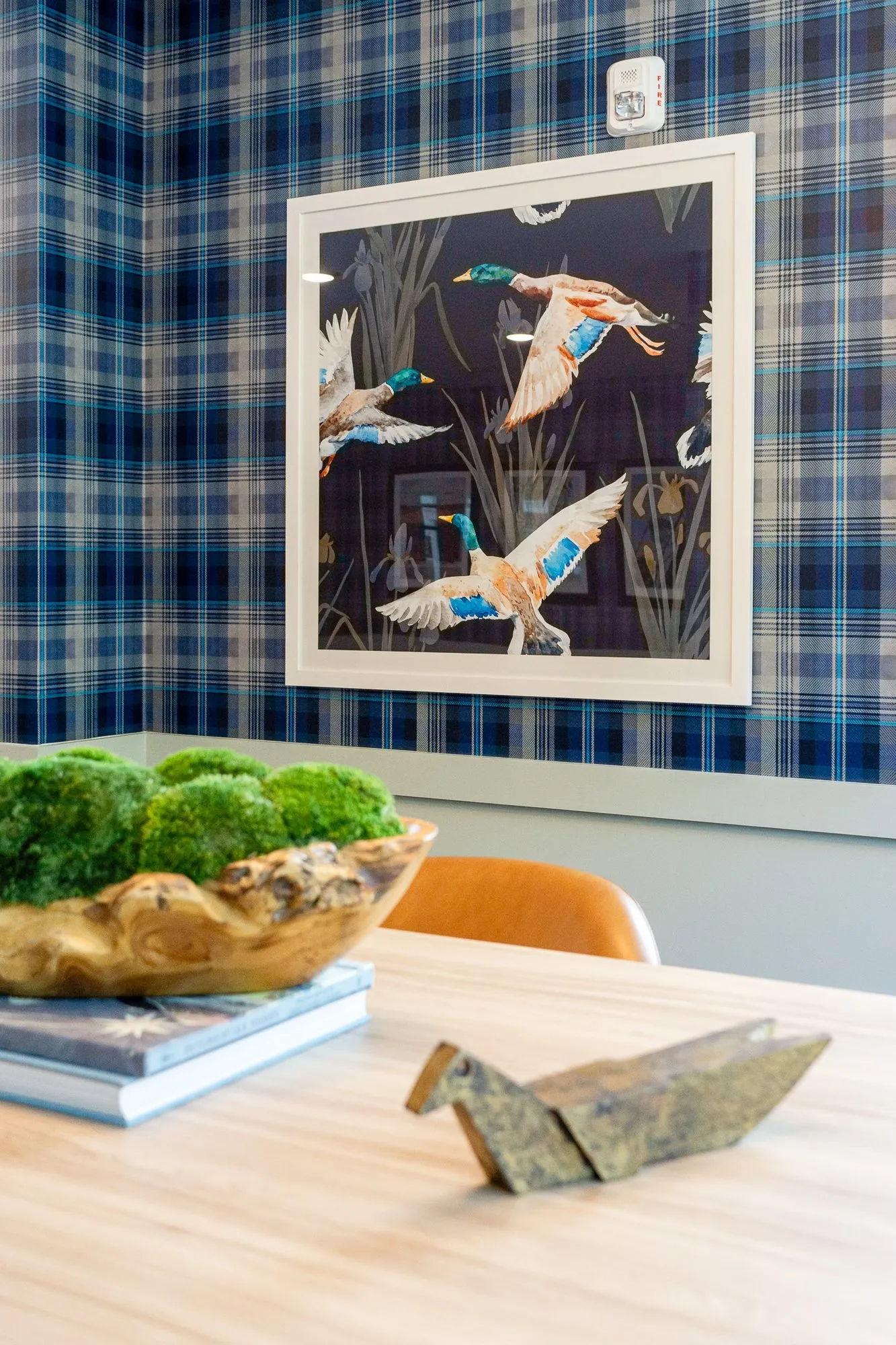 Multifamily amenity interior detail: framed mallard print on plaid wallpaper above a table with a moss bowl and brass bird