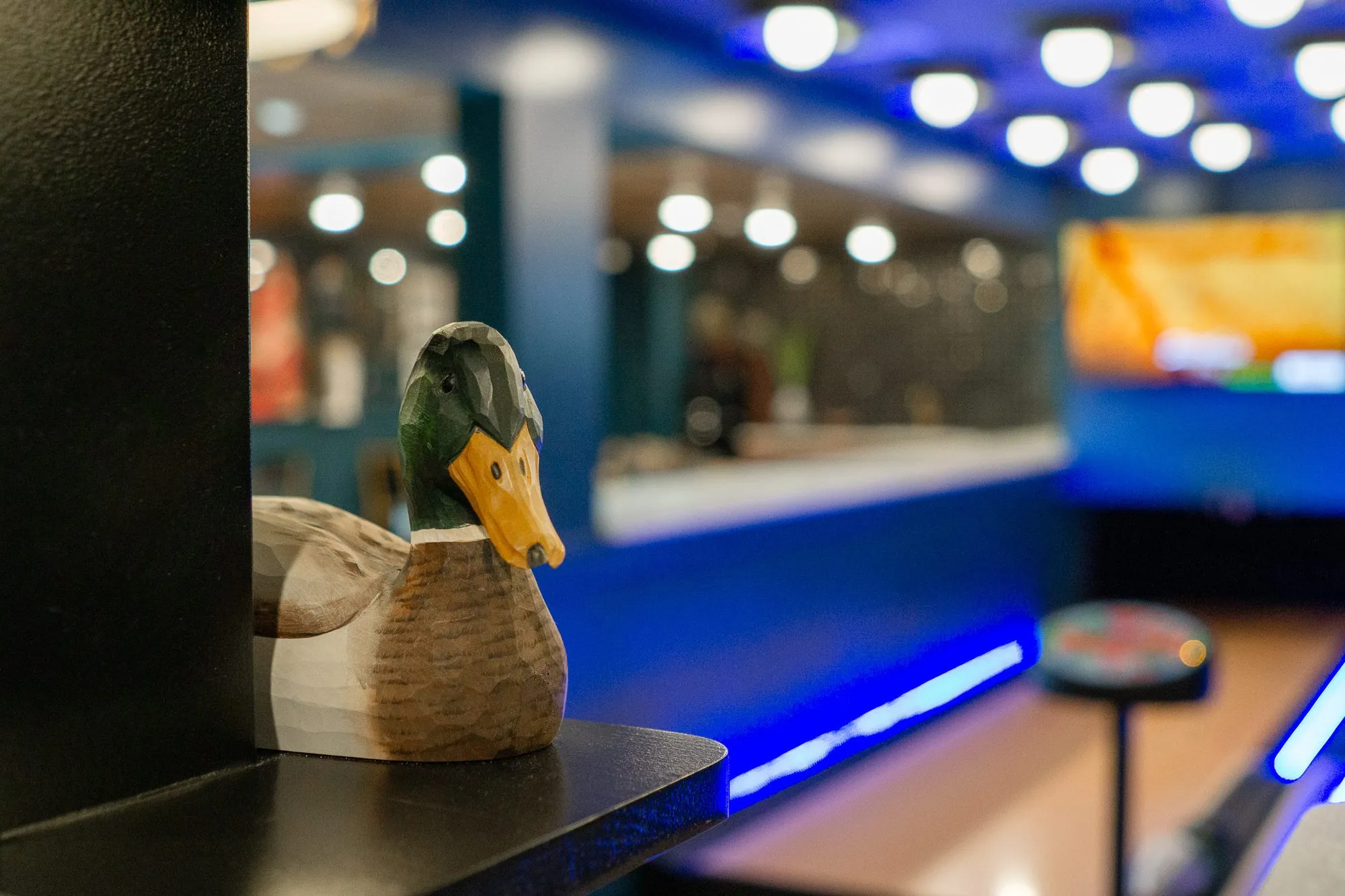 Multifamily amenity bowling lounge with a carved mallard decoy on a black shelf, the blue-lit lanes blurred beyond