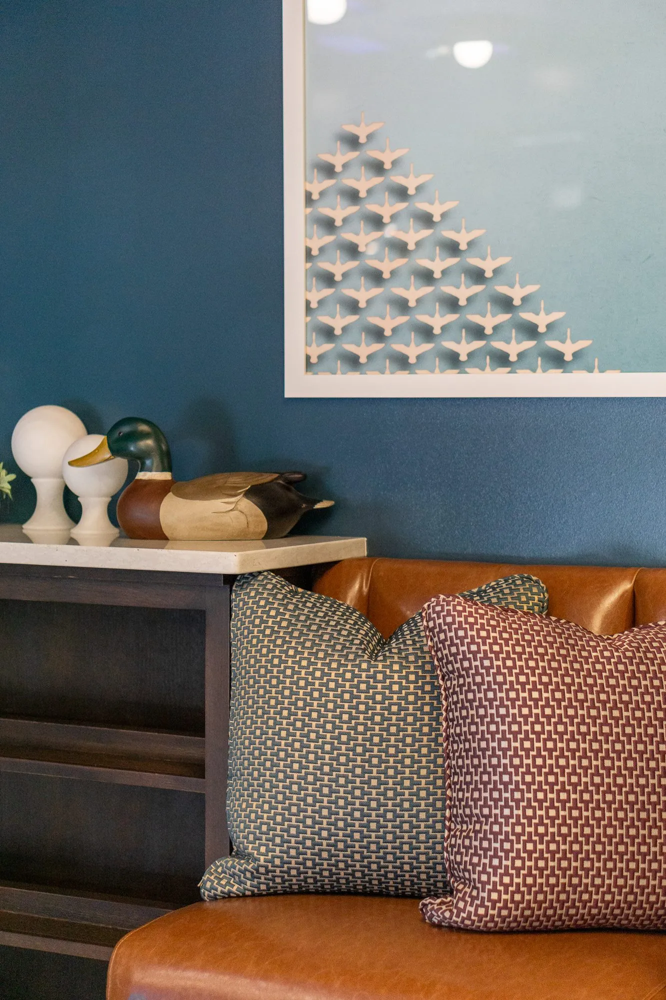 Multifamily amenity interior detail: cognac banquette with patterned pillows below a framed bird artwork, a bust lamp