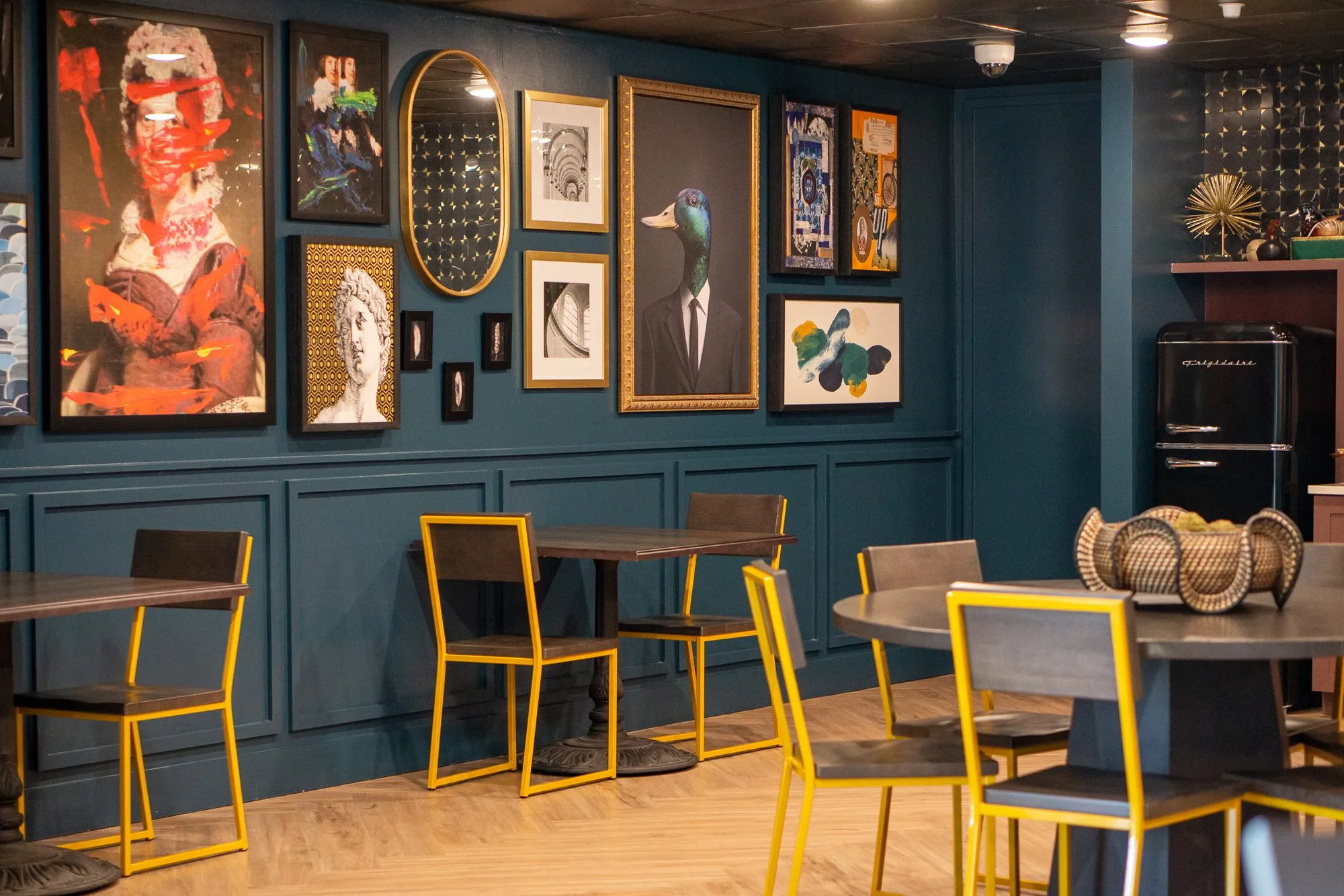 Multifamily amenity cafe with the teal gallery wall and yellow-framed chairs, a woven bowl on the round table