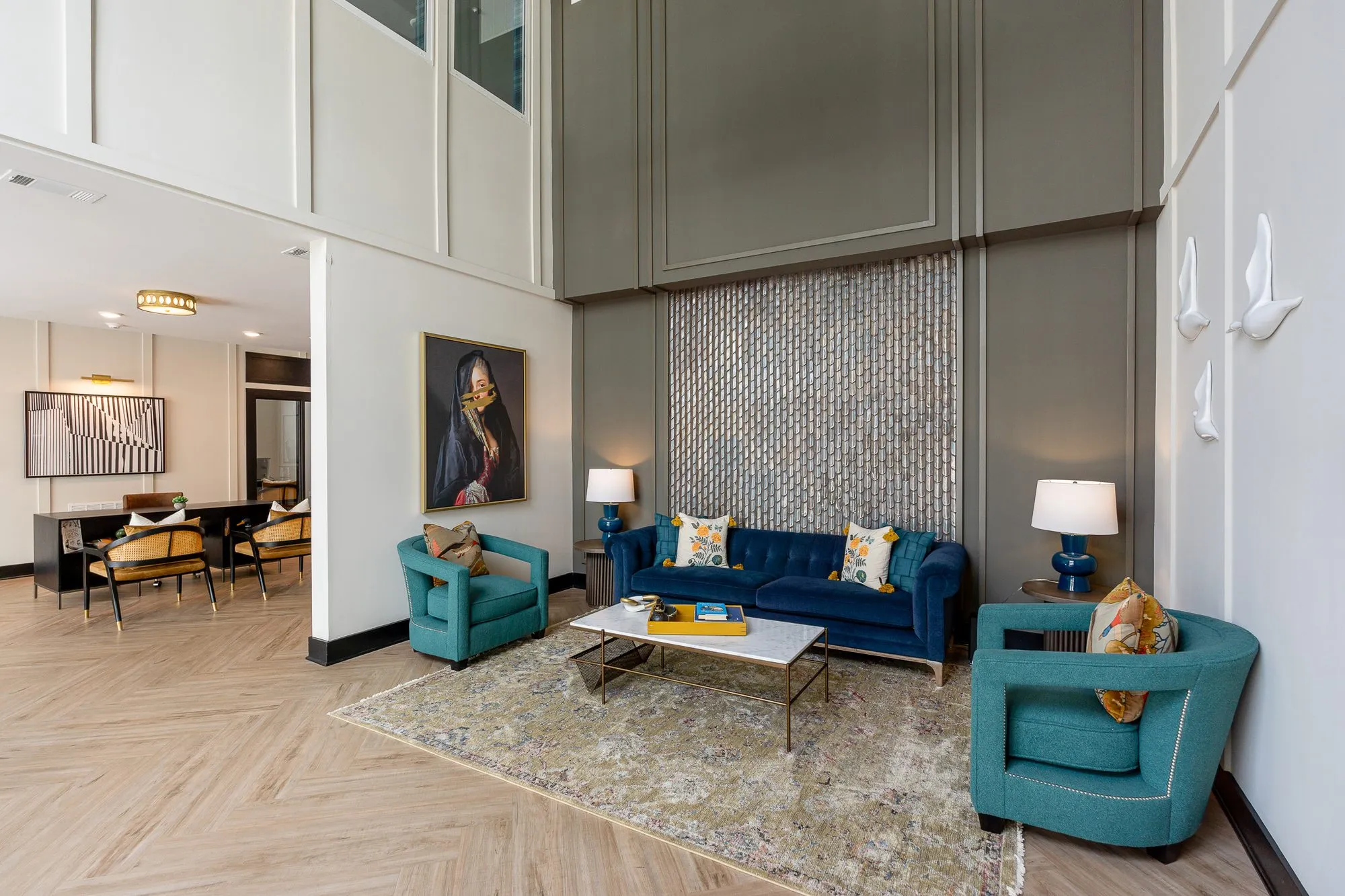 Multifamily amenity lobby lounge with a navy sofa and teal chairs below a tall tile panel, the leasing desk beyond