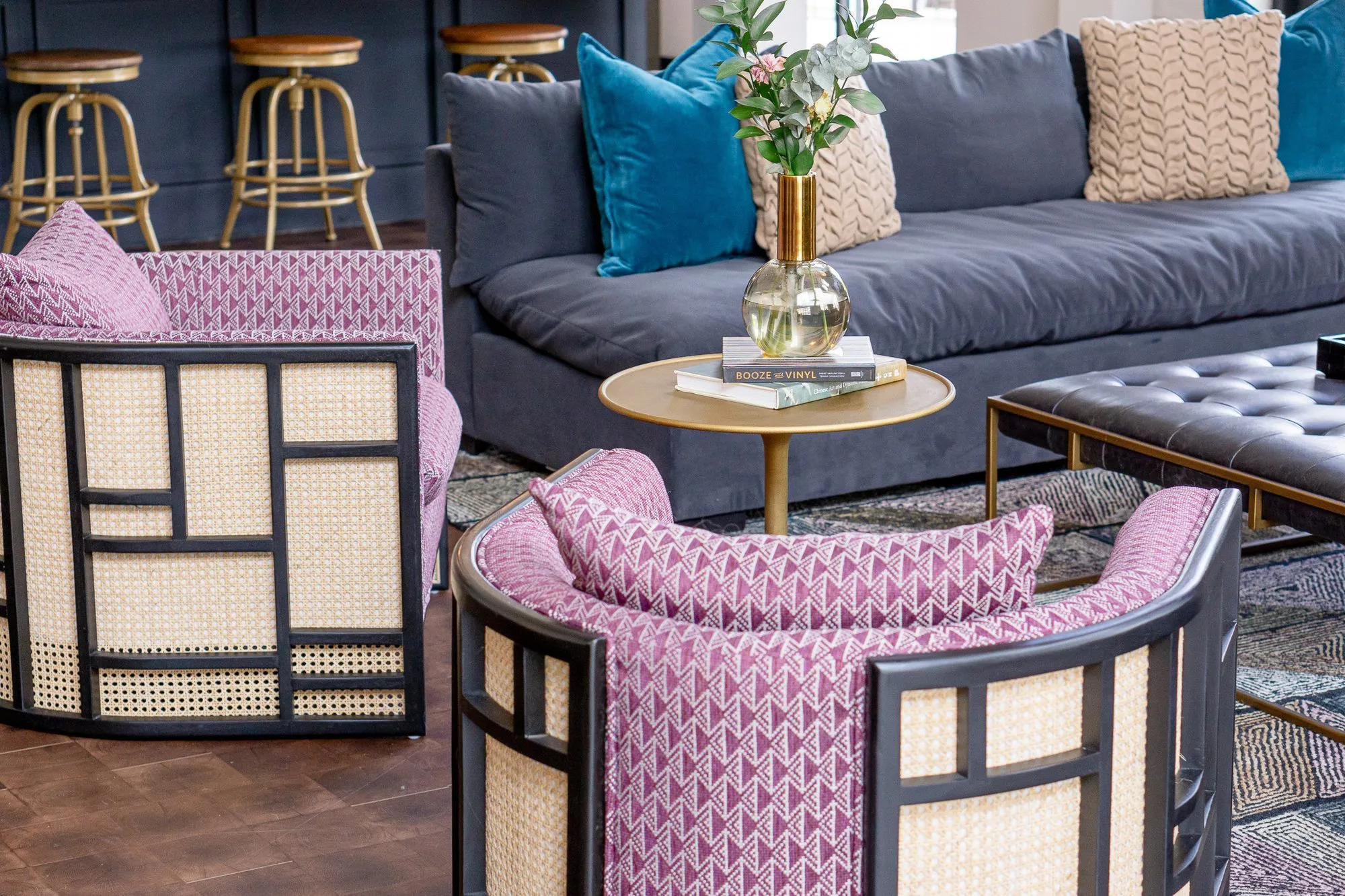 Multifamily amenity clubroom with pink patterned cane armchairs around a brass table, a navy sofa and the bar beyond