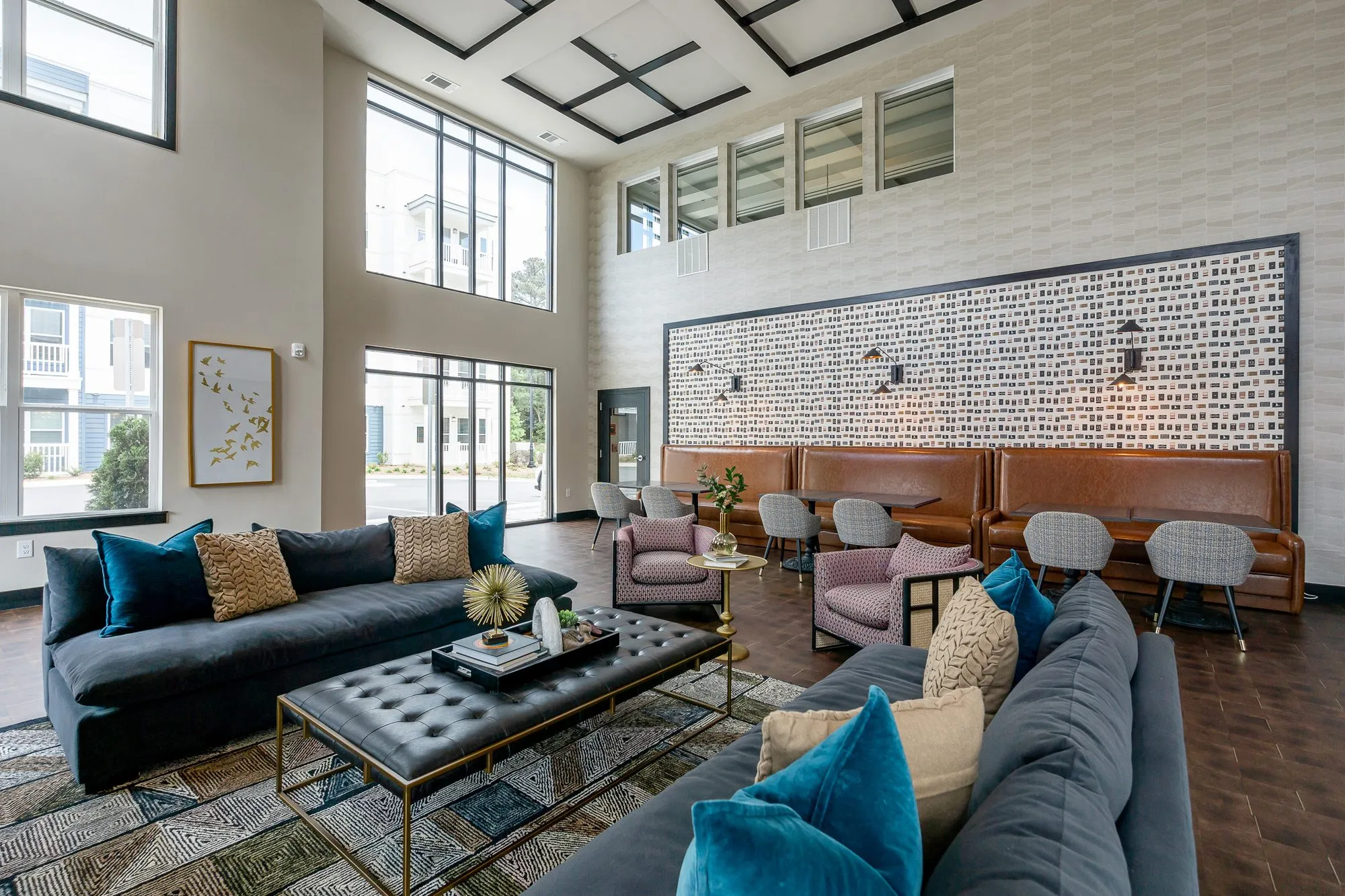Multifamily amenity clubroom with tall windows, navy sofas around a tufted ottoman and booths along the matchbook wallpaper