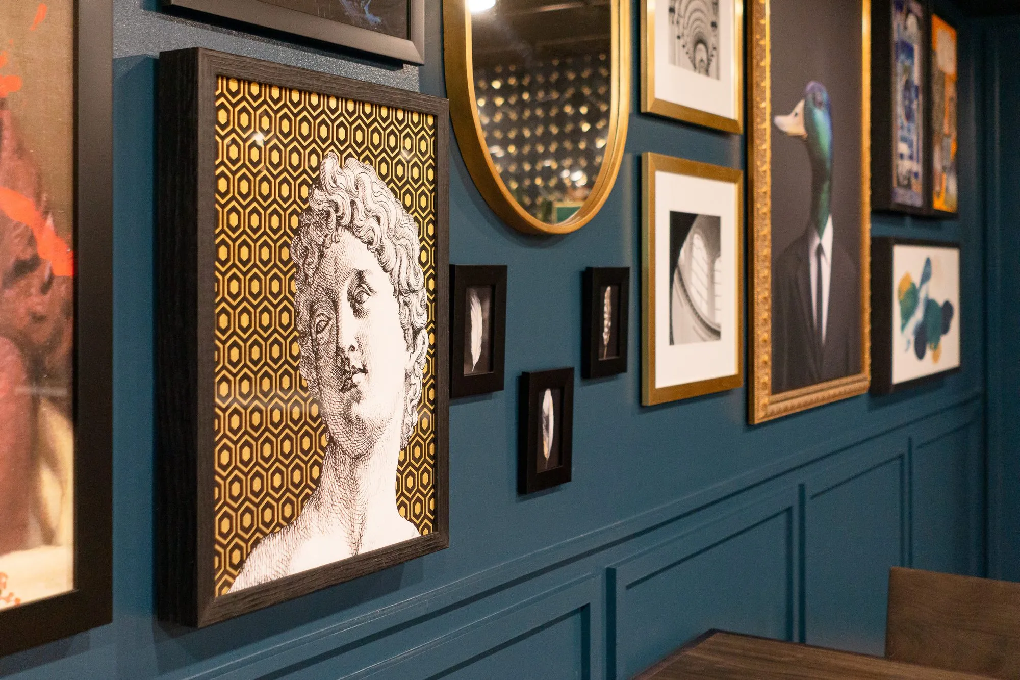 Multifamily amenity gallery wall on teal panelling with a bust print on gold hexagons, an oval mirror and the duck portrait
