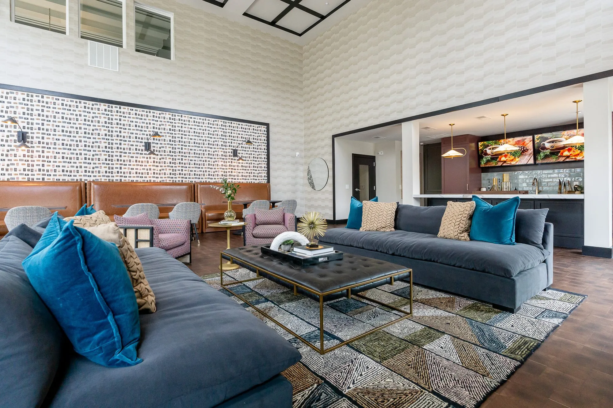 Multifamily amenity clubroom with a navy sofa and teal velvet pillows in front, a tufted ottoman and the bar beyond
