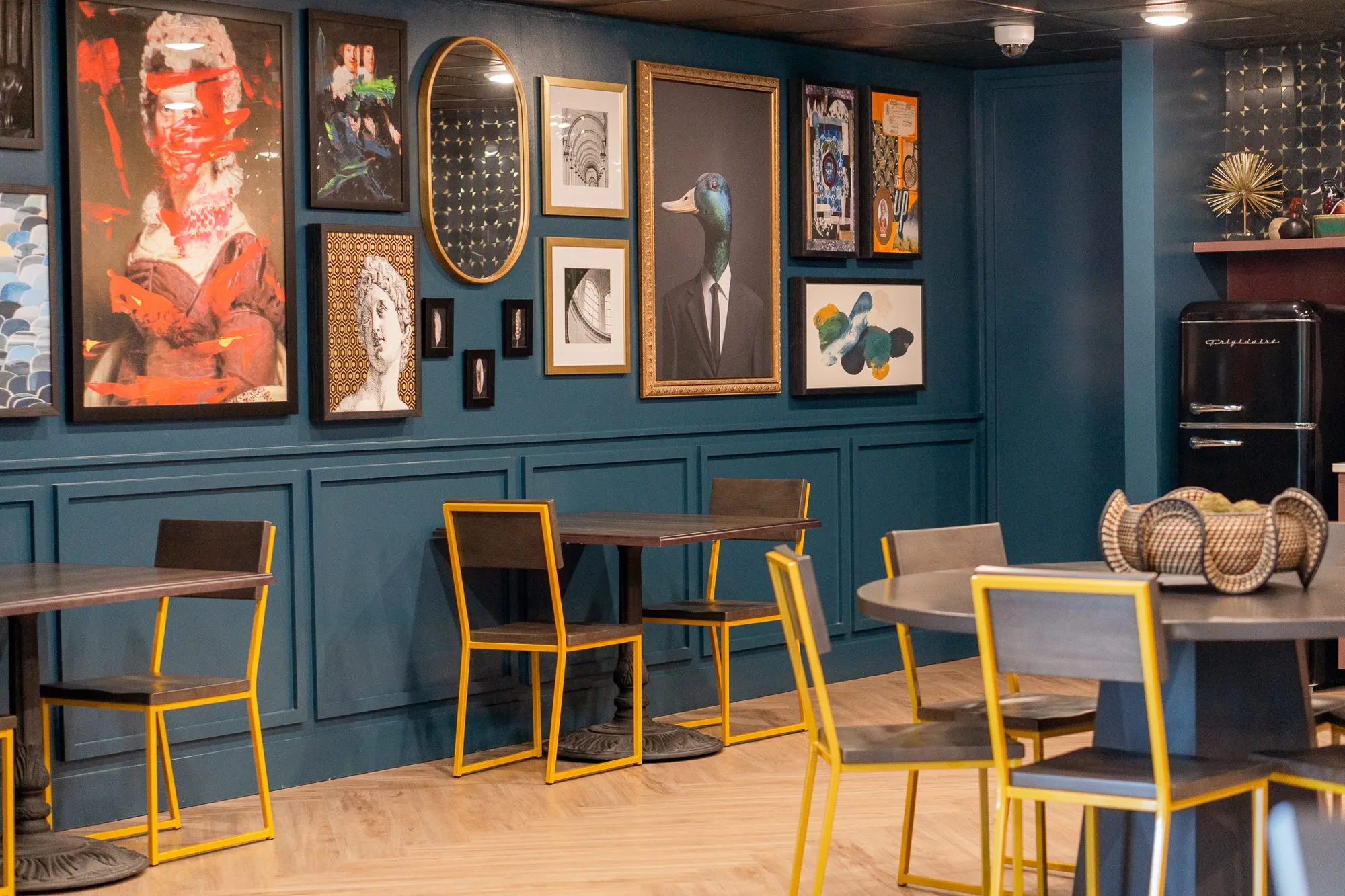 Multifamily amenity cafe with a teal gallery wall above yellow-framed chairs and dark tables, a retro refrigerator