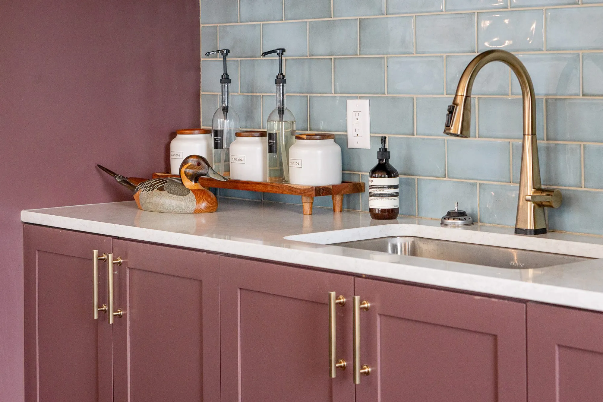 Multifamily amenity bar with mauve cabinets and brass pulls below a pale blue tile backsplash, a duck decoy on the counter