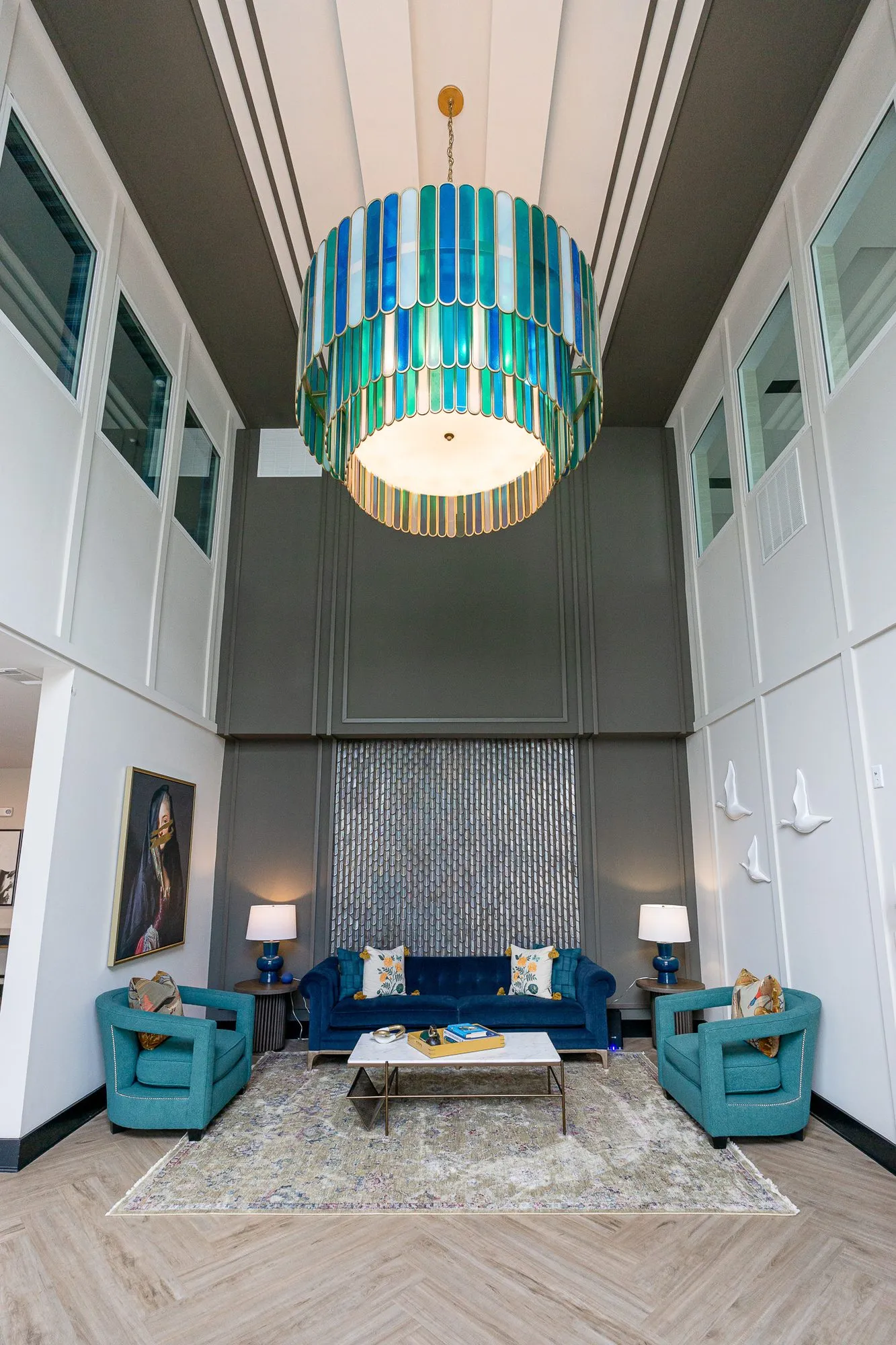 Multifamily amenity double-height lobby with the glass chandelier above a navy sofa and teal chairs