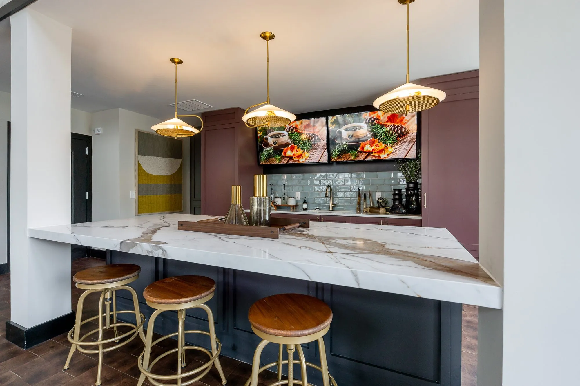 Multifamily amenity bar with brass pendants over a marble island and wood-seat stools, food screens on the mauve wall