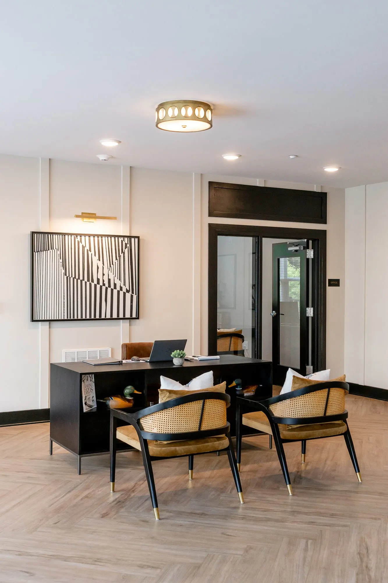 Multifamily amenity leasing area with a black desk and cane chairs below a stripe abstract, glazed doors behind