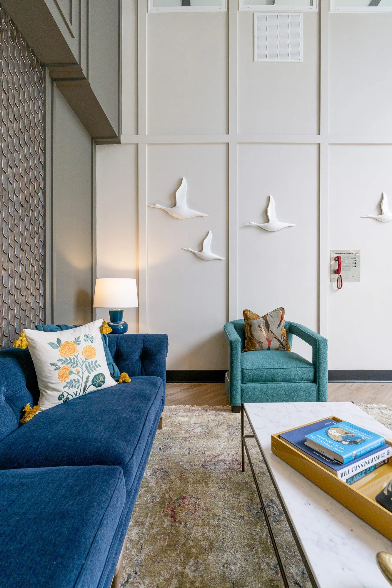 Multifamily amenity lobby lounge along a navy sofa with a floral pillow toward a teal armchair and white bird sculptures