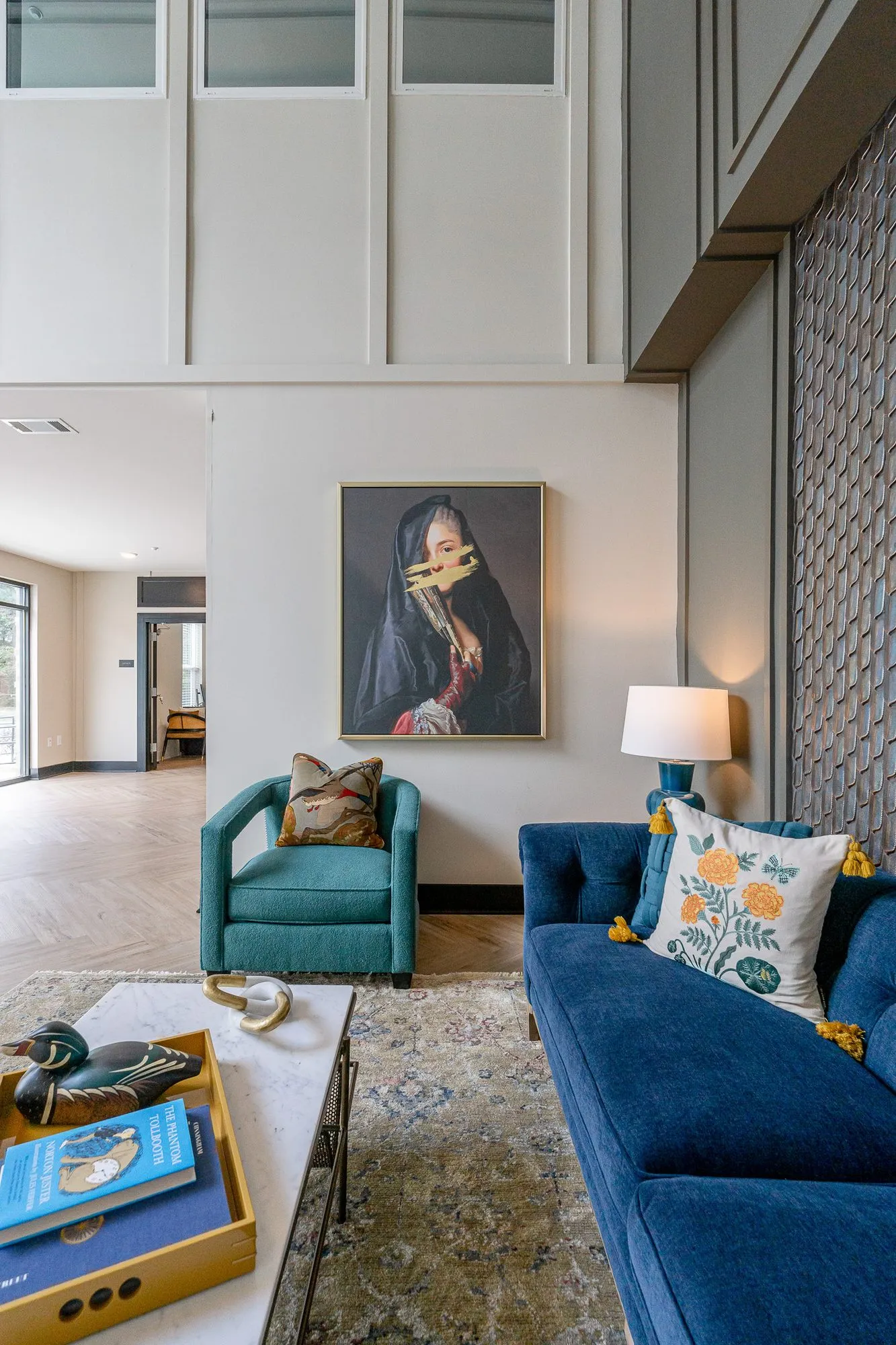 Multifamily amenity lobby lounge with a navy sofa and teal chair below the veiled portrait, a duck decoy on the tray
