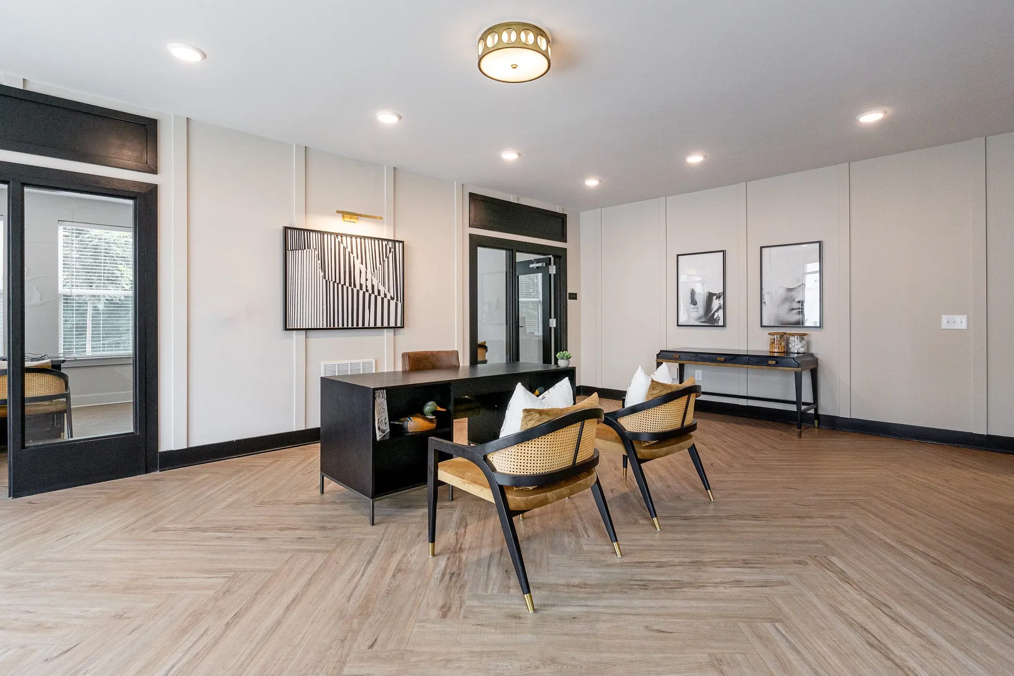 Multifamily amenity leasing area with a black desk and cane chairs on herringbone flooring, glazed doors and framed prints