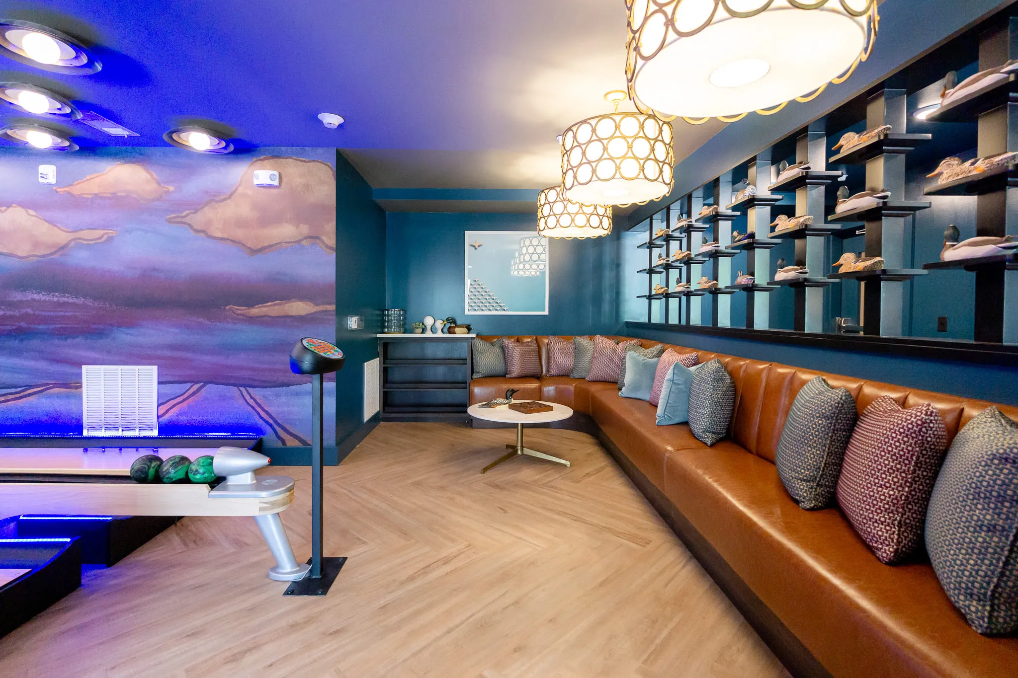Multifamily amenity duck-pin bowling lounge with a leather banquette and dark shelving beside the lanes under a marsh mural