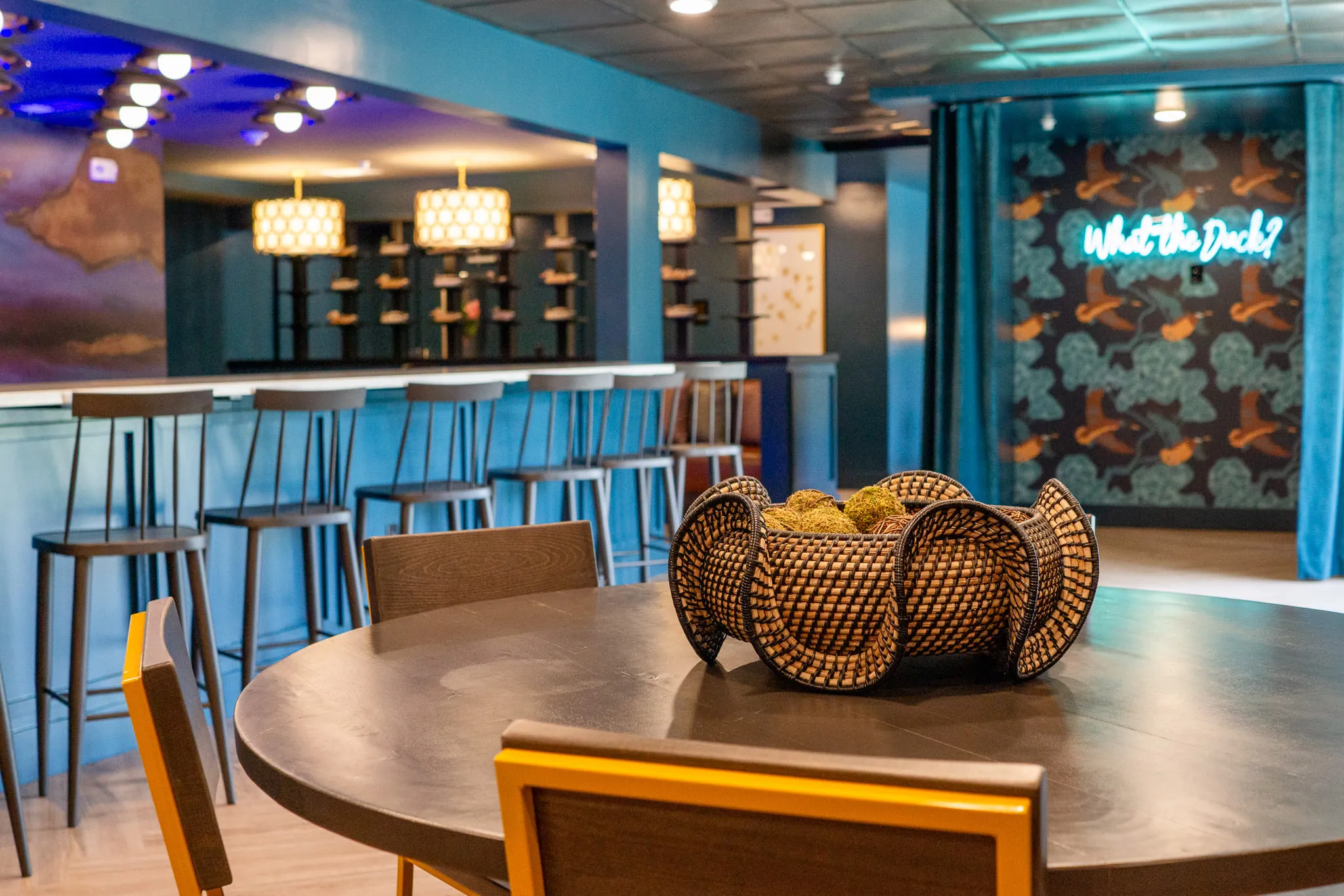 Multifamily amenity lounge bar with counter stools under pendant lights, a round wood table and the neon sign beyond