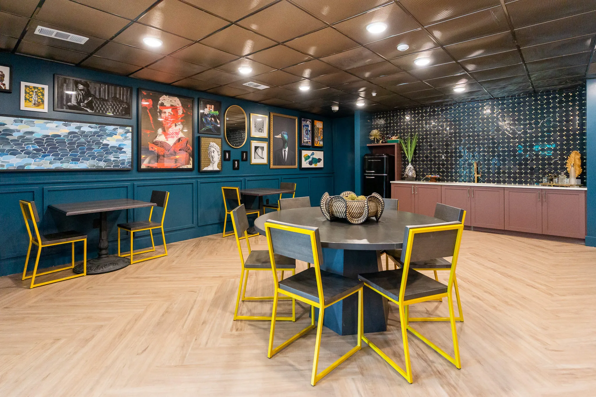 Multifamily amenity clubroom with yellow chairs and dark tables below a pressed tin ceiling and a dense gallery wall