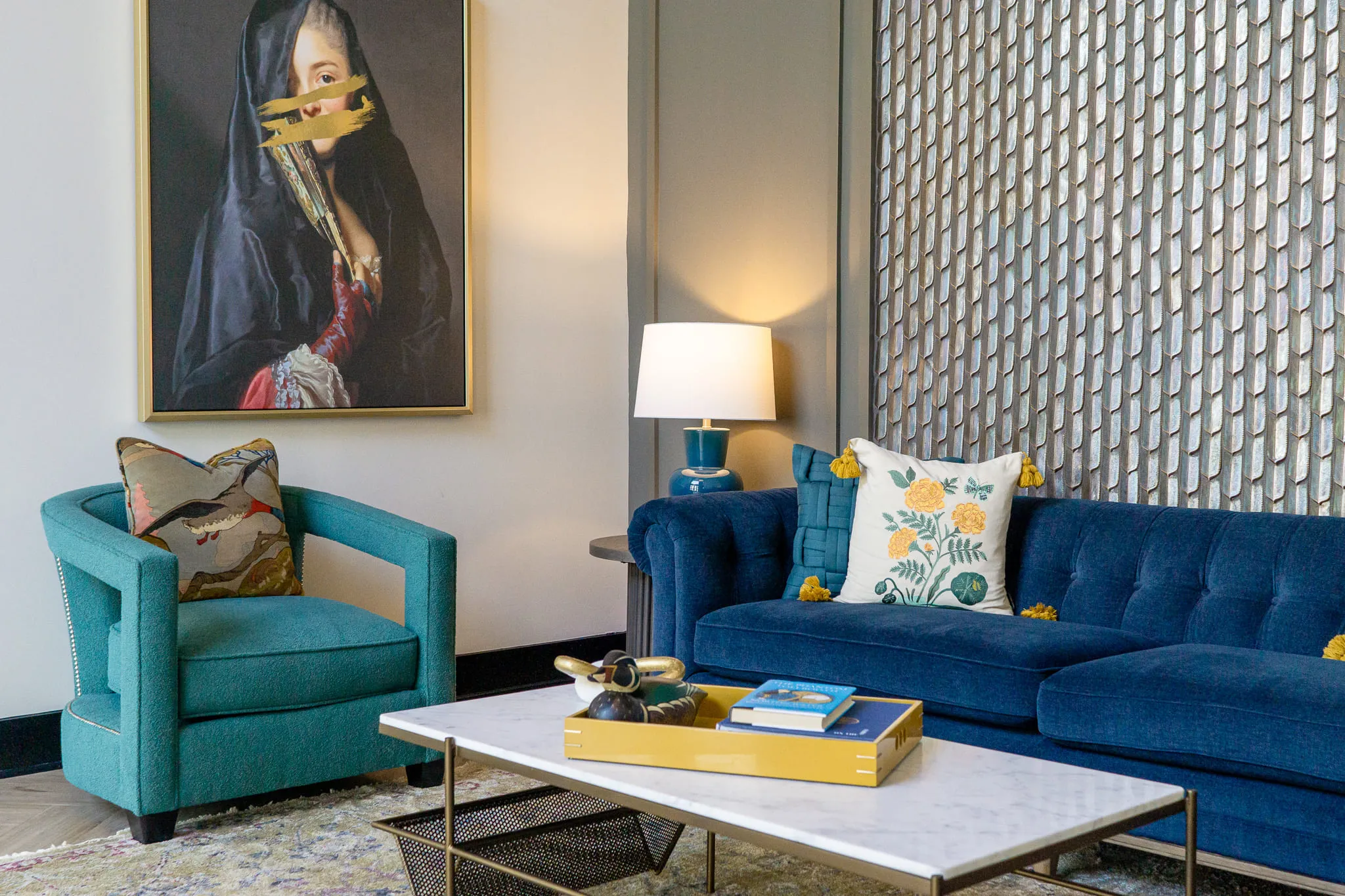 Multifamily amenity lobby lounge with a blue velvet sofa and teal armchair below the veiled portrait, a textured tile panel