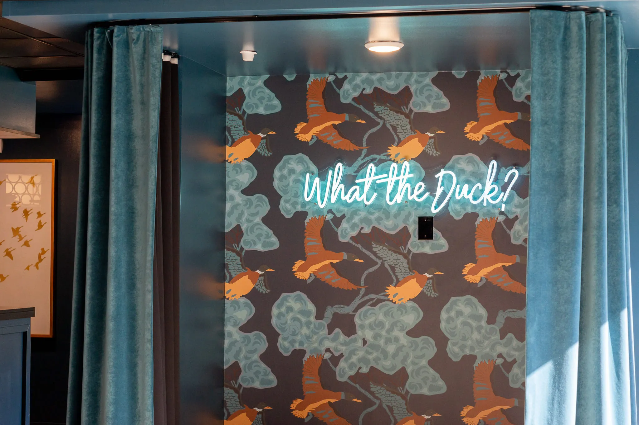 Multifamily amenity alcove with a neon sign reading What the Duck, on flying duck wallcovering between teal velvet curtains
