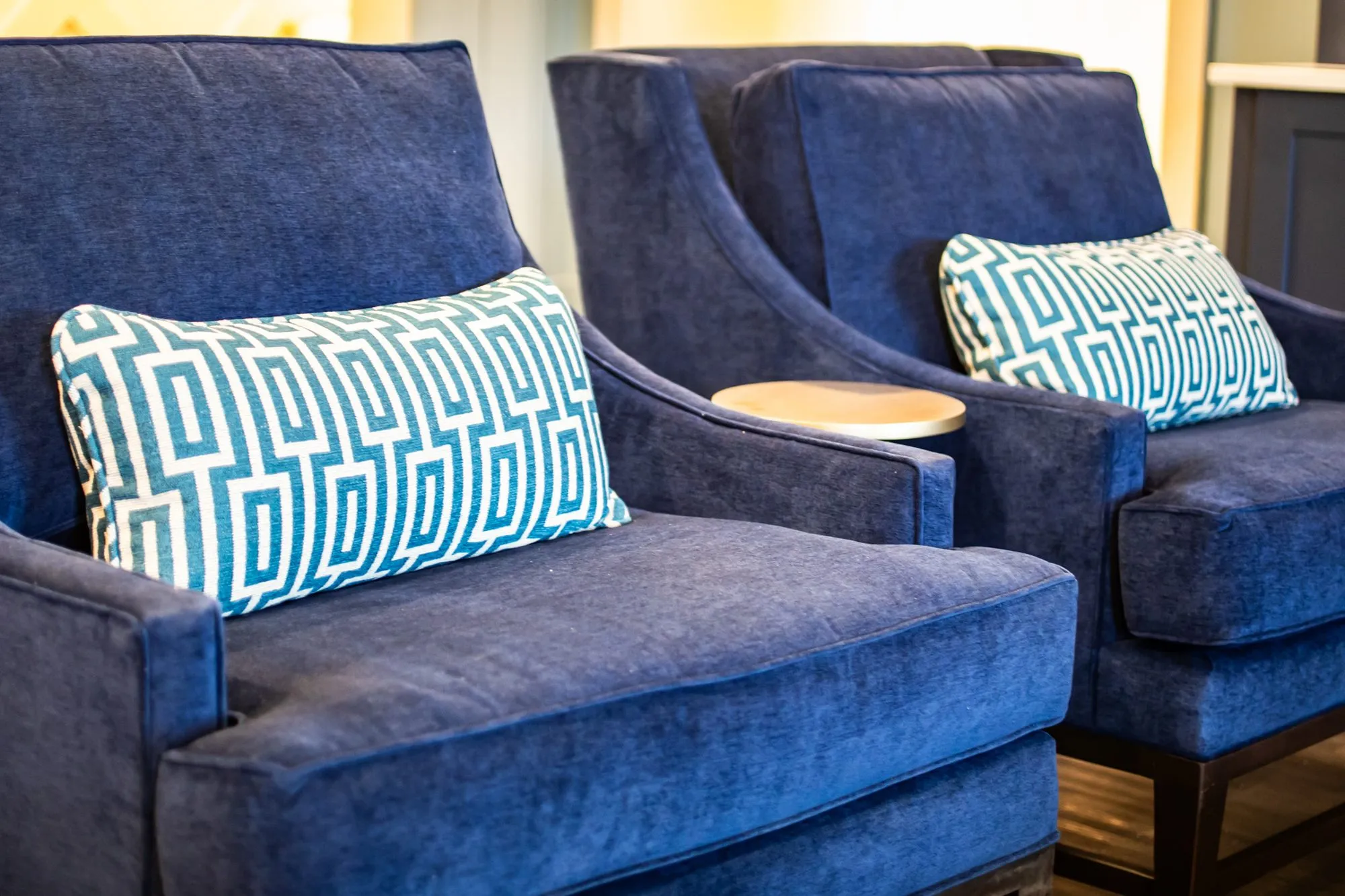 Senior living lounge with two navy velvet armchairs, teal Greek key lumbar pillows and a round side table
