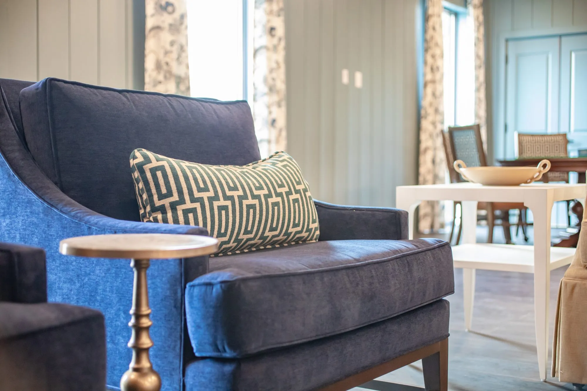 Senior living interior detail: navy velvet armchair with a Greek key pillow beside a brass side table, dining tables beyond