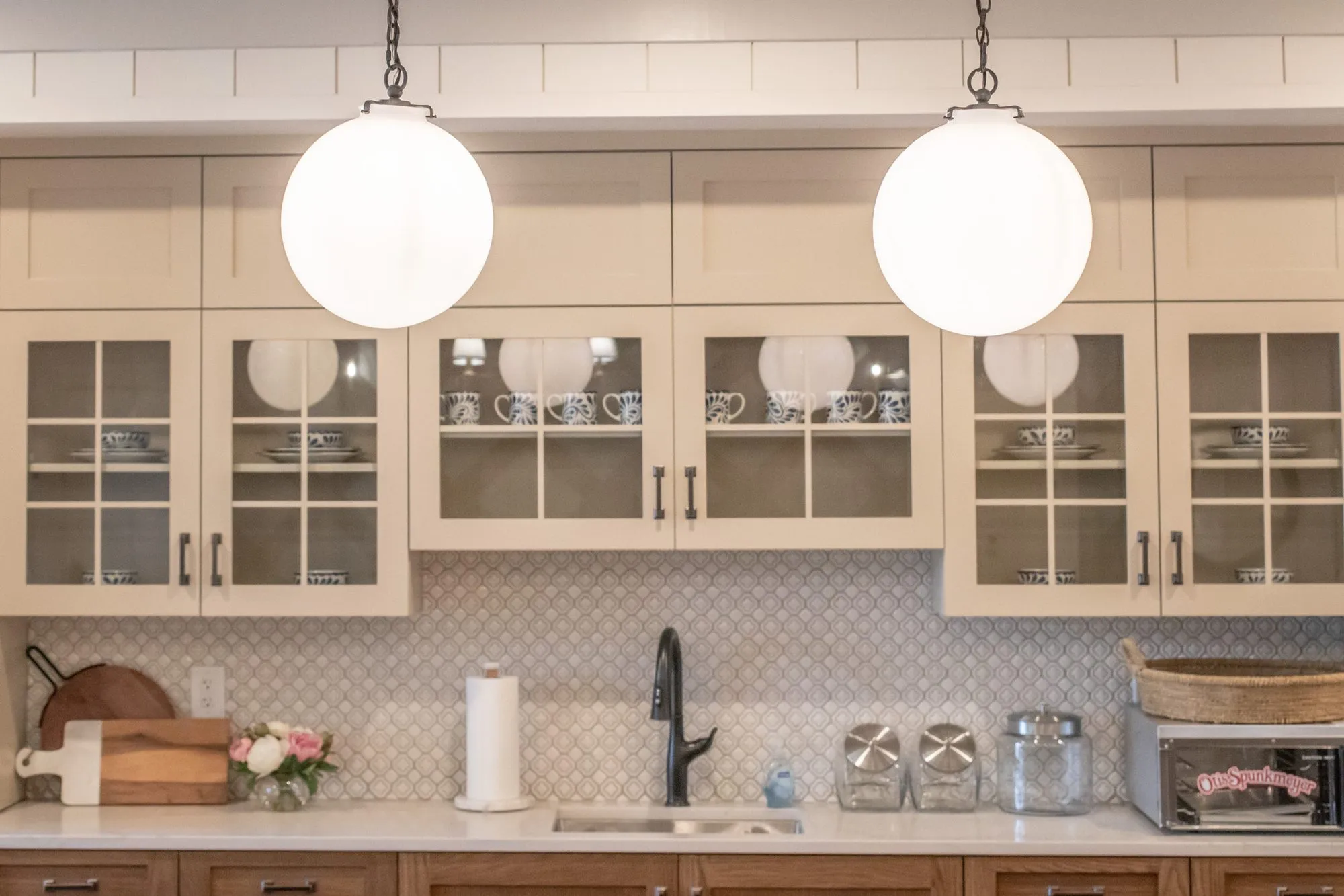 Senior living kitchenette with glass front cream cabinets, white globe pendants and a white arabesque tile backsplash