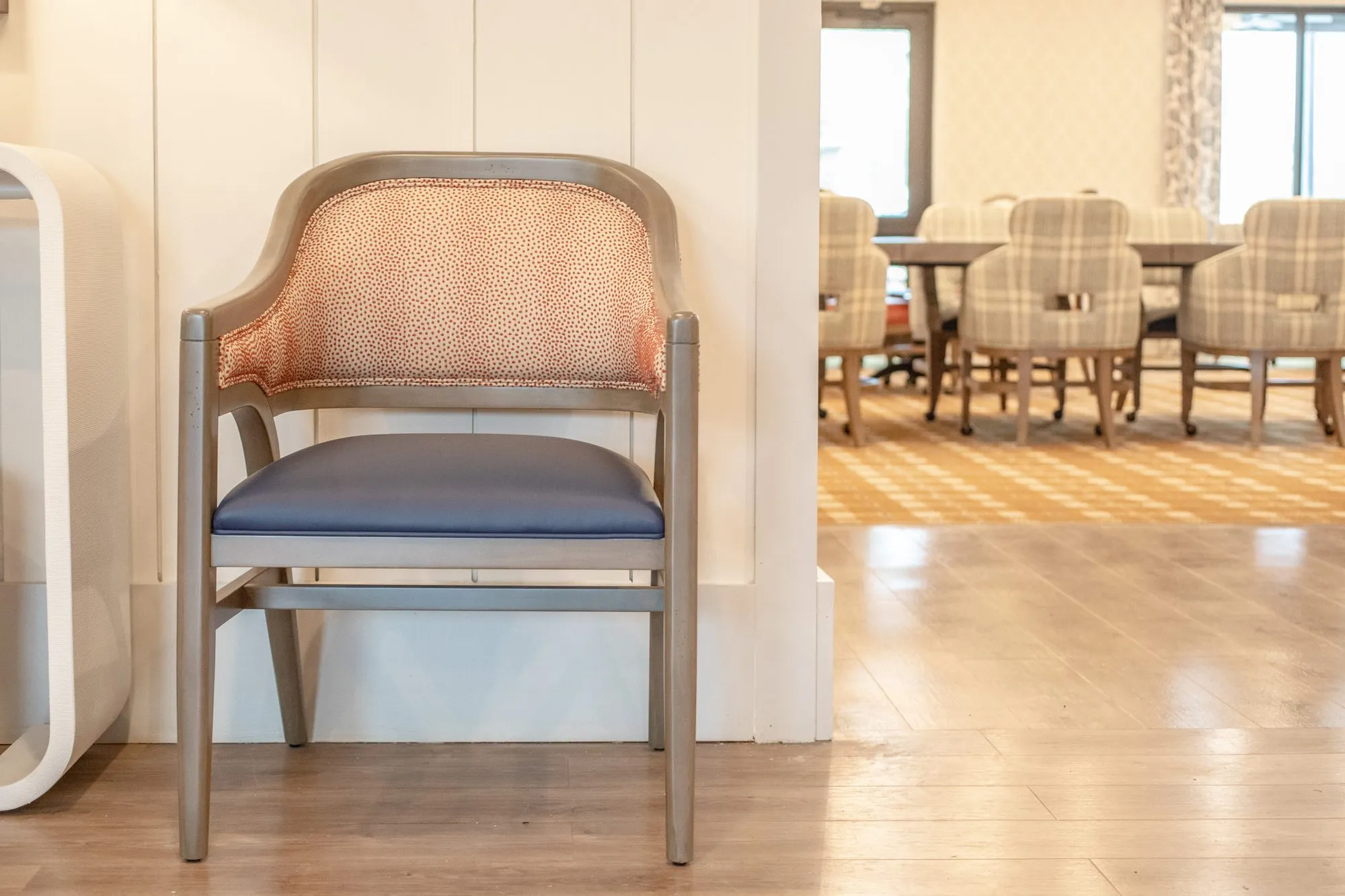 Senior living dining room: grey wood armchair with a coral dot back and navy seat, plaid chairs beyond