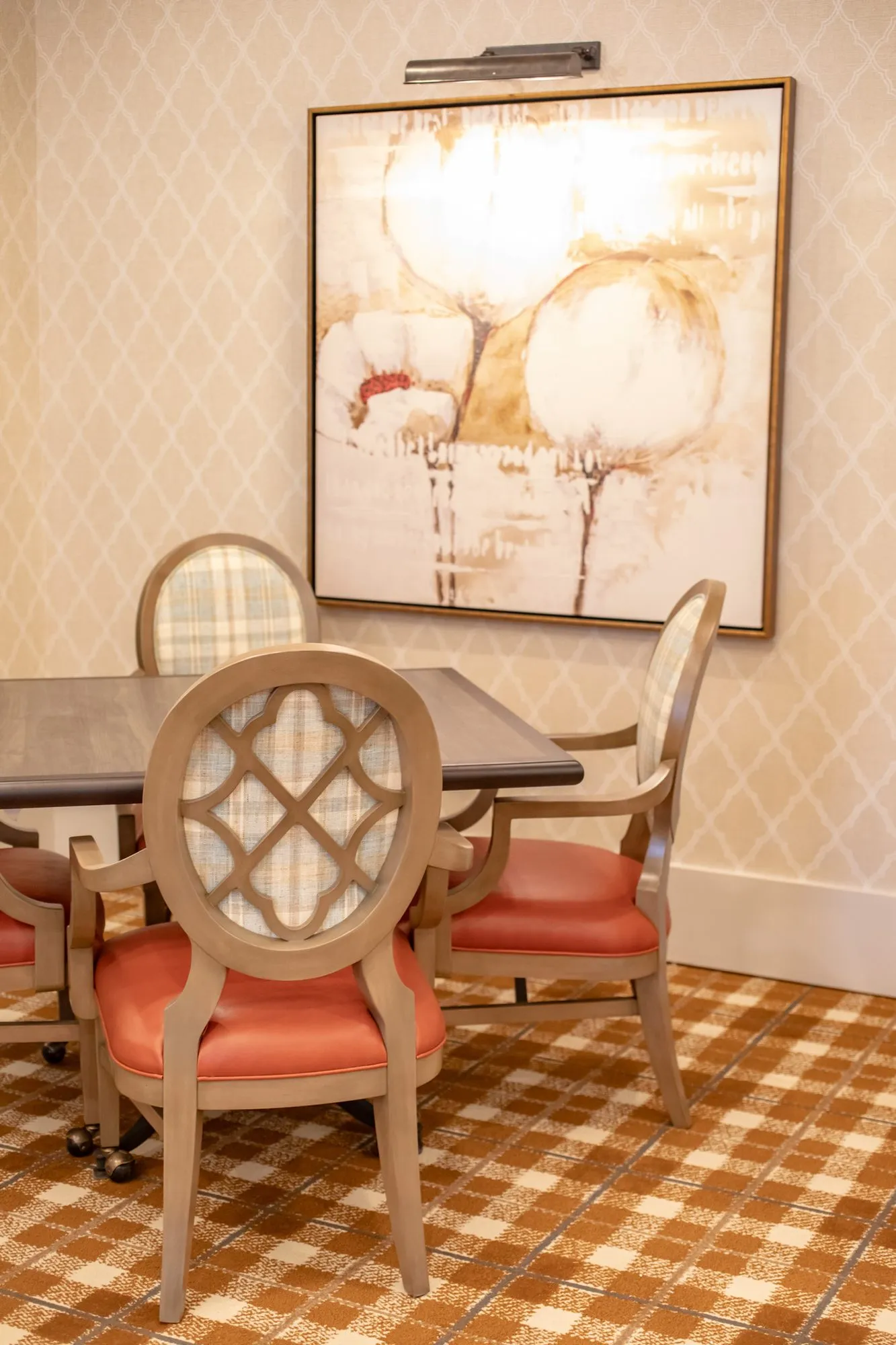 Senior living dining room with coral seated chairs at a square table below a cotton boll painting on trellis wallpaper