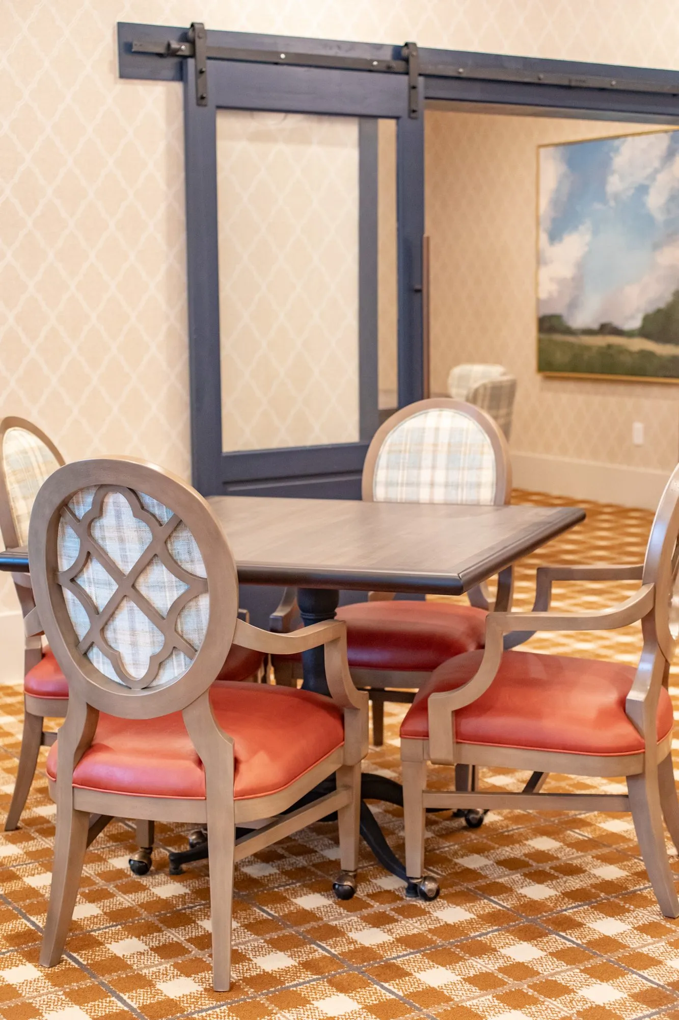 Senior living dining room with quatrefoil back chairs and coral seats at square tables, a navy barn door and landscape
