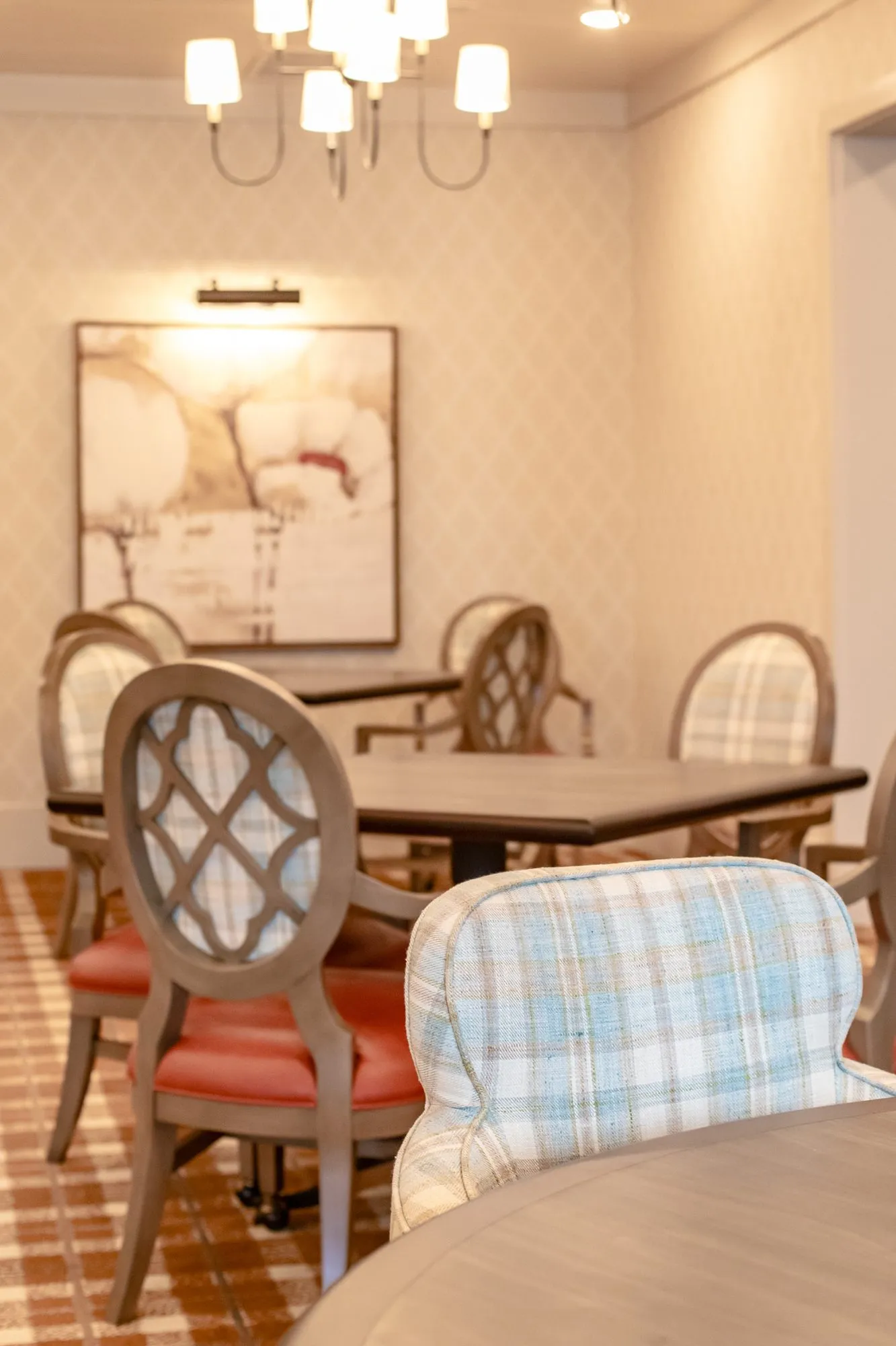 Senior living dining room with quatrefoil back chairs at square tables under a chandelier, a plaid chair in the foreground