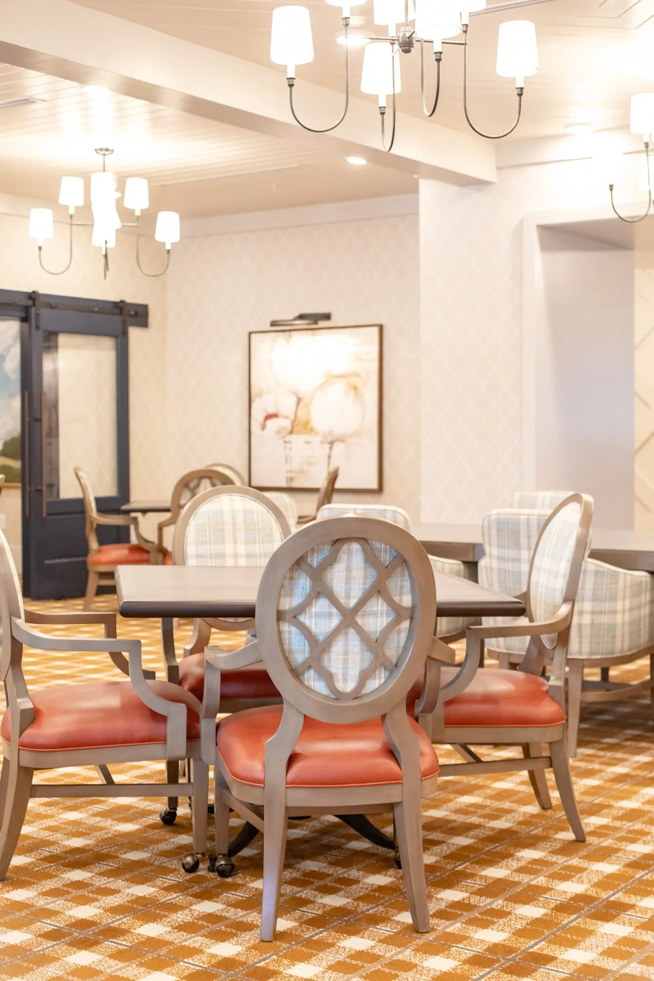 Senior living dining room with candle chandeliers above coral seated chairs at square tables and a barn door