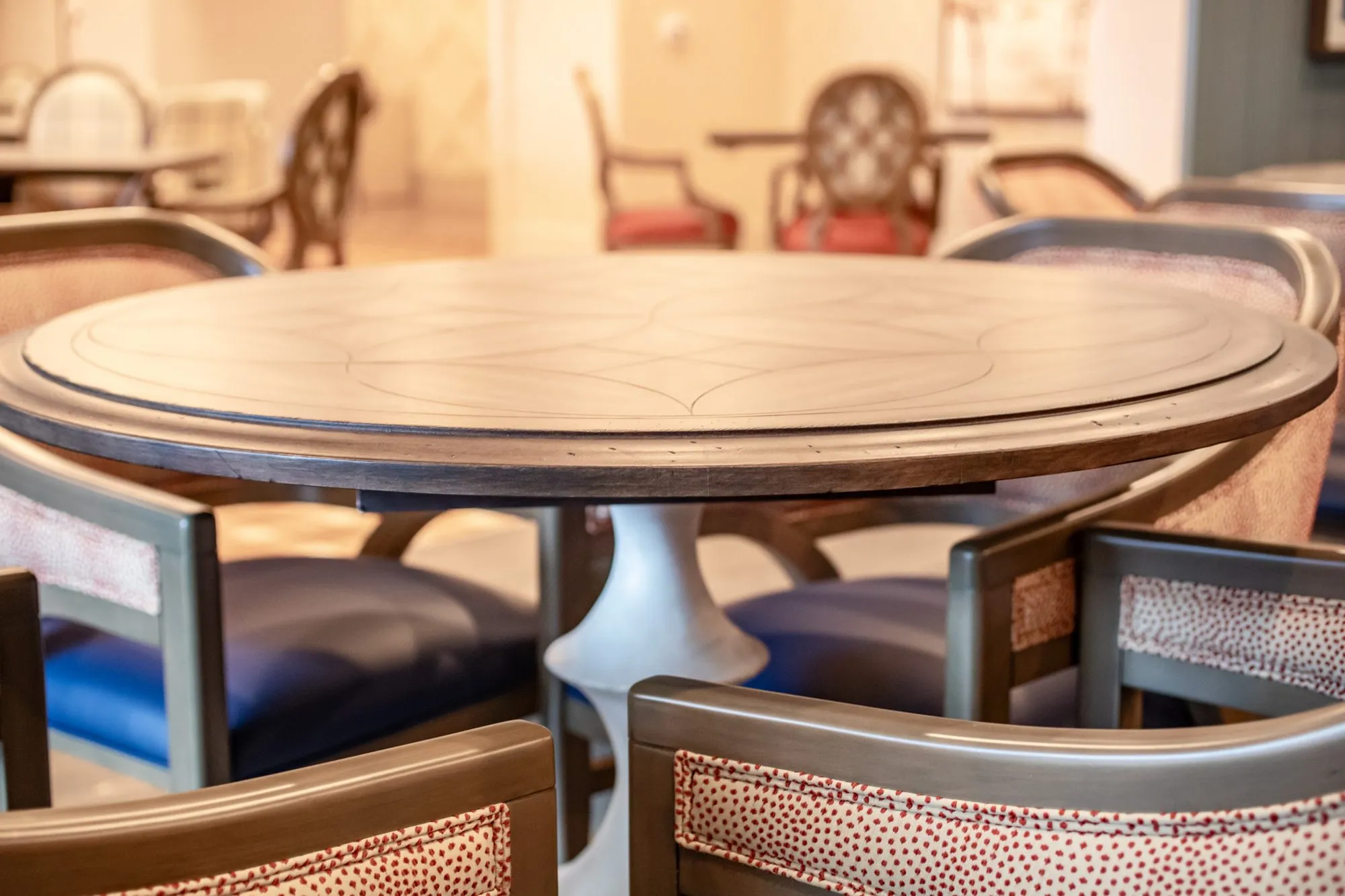 Senior living interior detail: round wood table with a flared pedestal base surrounded by coral dot and navy chairs