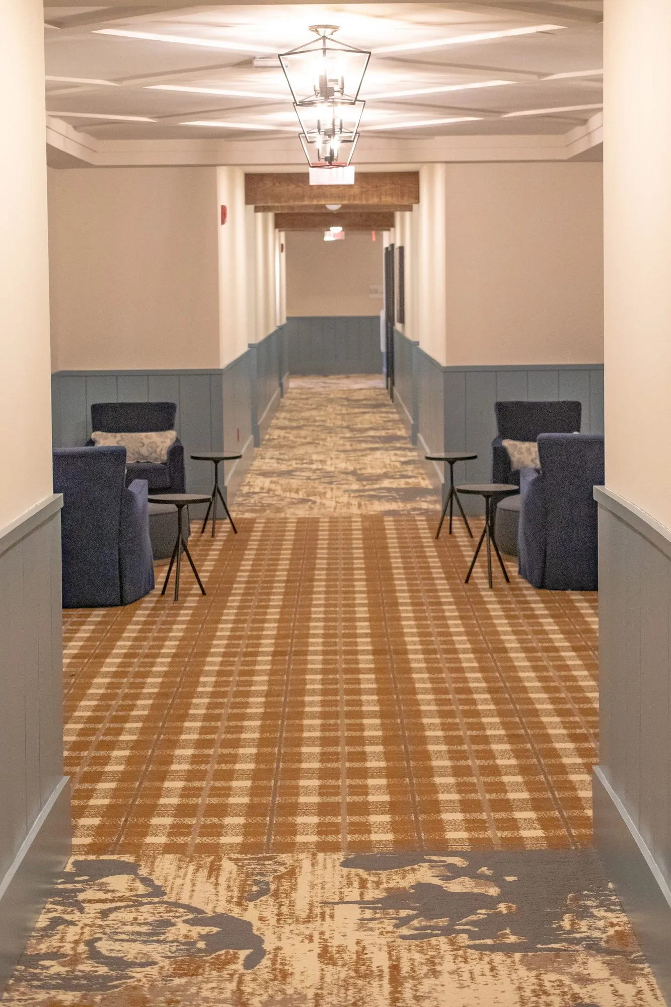 Senior living corridor with plaid carpet, navy armchairs either side and lantern pendants under a coffered ceiling